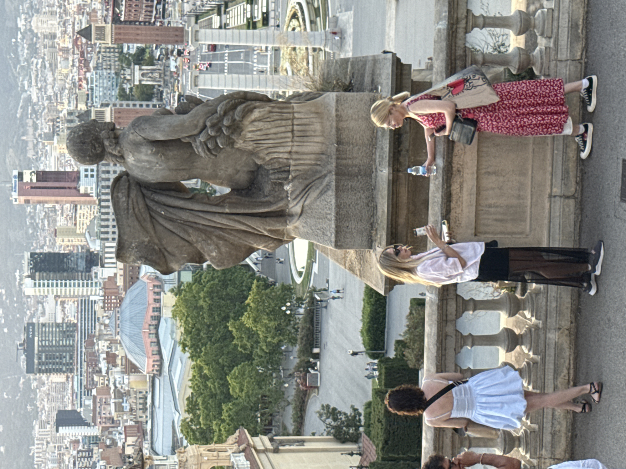 The image depicts a scenic viewpoint overlooking the city of Barcelona, Spain. In the foreground, three individuals are visible on a stone balcony. On the left, a person in a light blue dress stands with their back to the camera, facing away from the city view. To their right, a woman with blonde hair, wearing a pink shirt and black skirt, holds a drink and converses with another blonde woman. This second woman, dressed in a red floral dress and white sneakers, is holding a water bottle and looking towards her companion. Behind the individuals, a large stone sculpture of a human figure is prominently displayed. Beyond the balcony, the cityscape unfolds with numerous buildings, including high-rises and a distinctive domed structure. Lush green trees are scattered throughout the middle ground, and a large plaza with pathways and people can be seen below. The background is dominated by the sprawling urban landscape of Barcelona, with distant mountains or hills completing the vista. The lighting suggests it is daytime, possibly late afternoon, with soft sunlight illuminating the scene.