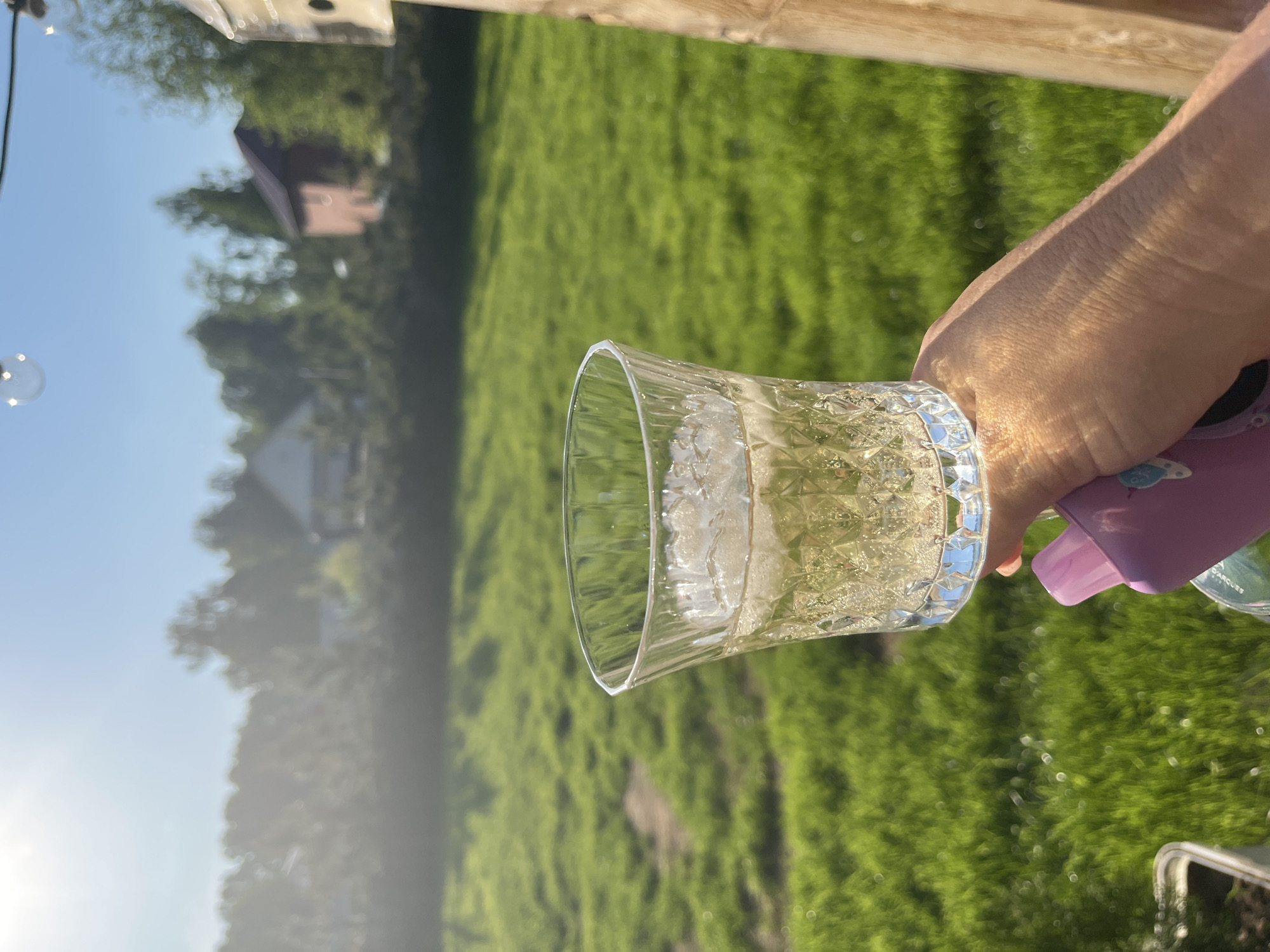 A hand holds a clear glass filled with a light-colored beverage and ice. The glass is patterned. Behind the hand and glass, a green lawn is visible. In the blurred background, there are trees and a building. A string of lights can be seen to the left. The location is not identifiable as Новая Чернушка, Russia from the provided image.