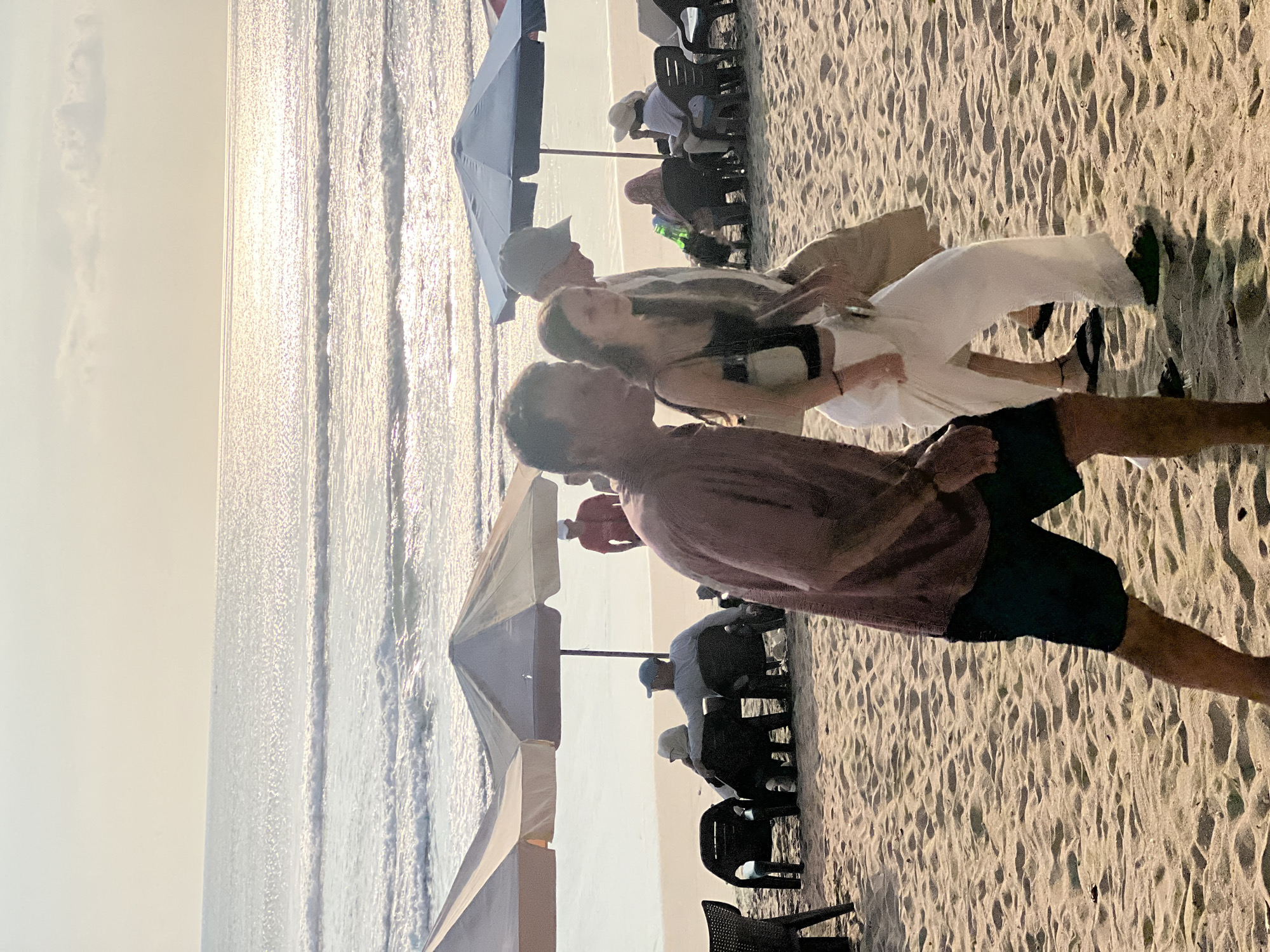 The image captures a lively scene on Kuta Beach, Indonesia, during what appears to be late afternoon or early evening. The sun is low in the sky, casting a warm, golden glow that reflects off the gentle waves of the ocean. The sand is rippled, indicating the tide has recently receded.
In the foreground, two people are walking on the beach. A man in a maroon short-sleeved shirt and black shorts is in profile, looking to his right. Next to him, a young woman in a black tank top and flowing white pants is also walking, her gaze directed forward. Behind them, a man wearing a light-colored cap and a tan jacket walks alongside the woman.
Further back, several beach umbrellas provide shade for seated individuals. One umbrella is dark blue, and another is cream-colored. People are seen sitting in black chairs, some facing the ocean, others conversing. A person in a red shirt stands near one of the umbrellas. The overall impression is one of a relaxing day at the beach with various activities taking place.