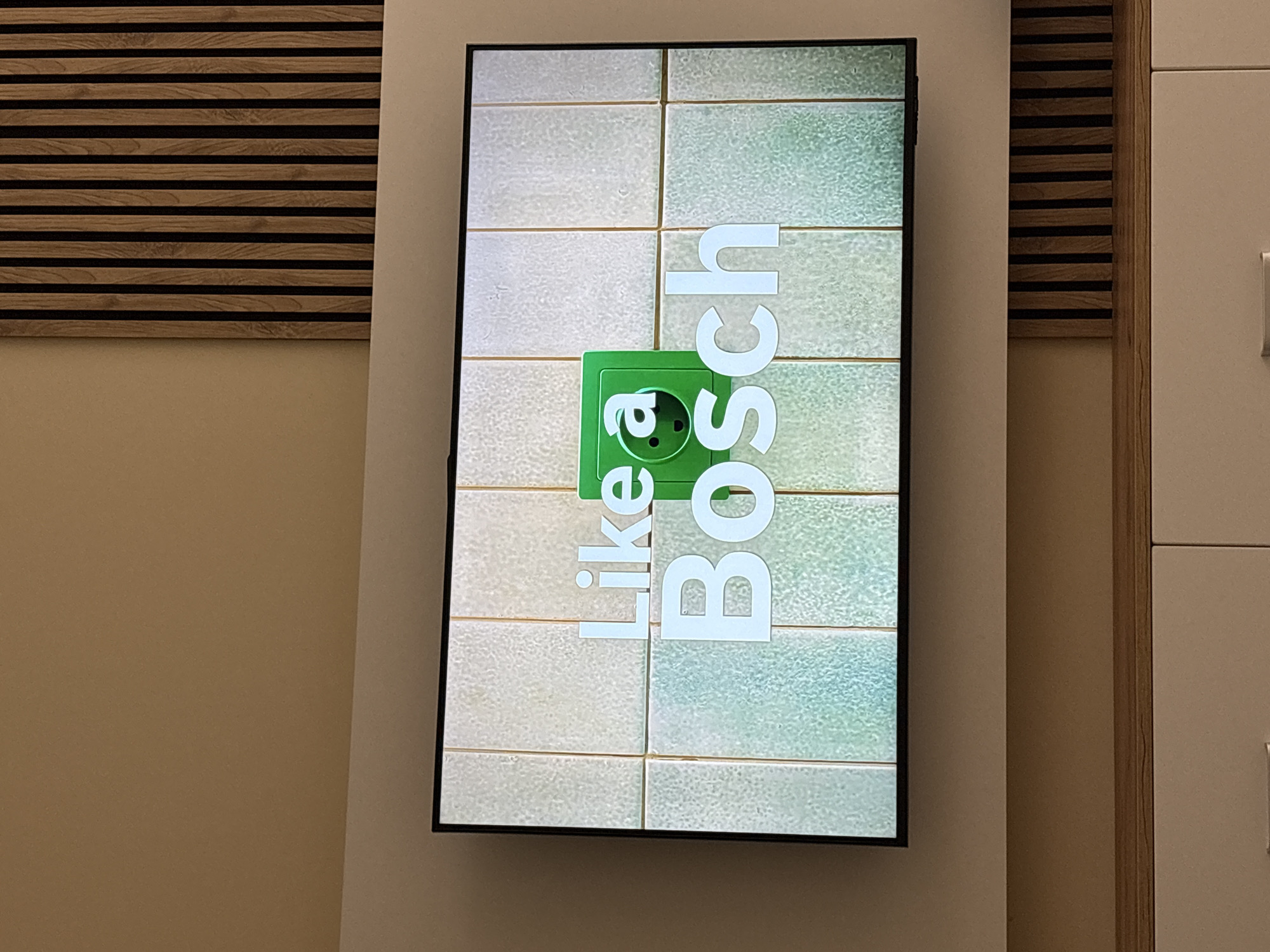 A vertically oriented digital screen displays an advertisement in an indoor setting. The screen's content features a tiled background, with the left side being light grey and the right side light blue-green. Centered on the screen is a bright green electrical socket symbol. Overlaid on this background, and rotated 90 degrees clockwise, is white text that reads "like a Bosch". "like a" is in a smaller font, while "Bosch" is in a larger, bolder sans-serif font.
The screen is integrated into a light beige or off-white wall panel. To the left of the screen, a decorative wall panel made of horizontal wooden slats on a black base is visible, above a plain light brown wall section. To the right of the screen, part of a light-colored cabinet or piece of furniture with a vertical wooden frame is seen, suggesting a modern interior. A glimpse of a silver handle on the far right indicates a door or drawer.
The lighting is consistent with an artificial indoor environment. No people are present. The scene depicts a commercial or exhibition space, likely a showroom, office, or public area, located in Sofia, Bulgaria, promoting the Bosch brand.