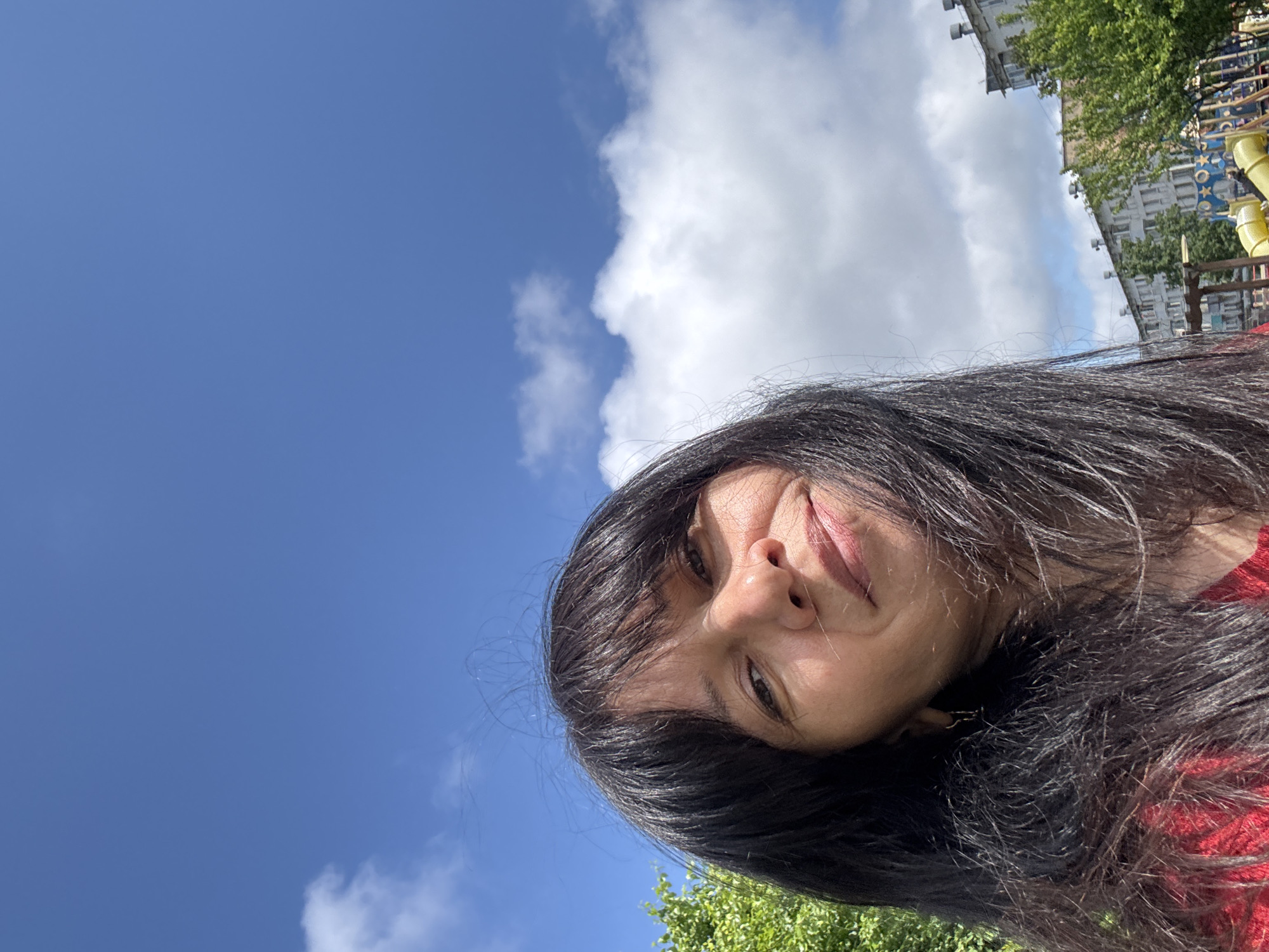 The image displays a bright, outdoor selfie of a woman in Chaikovsky, Russia, taken on a clear, sunny day. The woman, with long, dark, straight hair framing her face, occupies the lower right portion of the frame. She has dark eyes, a small nose, and her lips are curved in a subtle, closed-mouth smile. A glimpse of red clothing is visible on her right shoulder. The majority of the background consists of a vibrant blue sky with scattered white, fluffy clouds, suggesting good weather. In the upper right corner, hints of an urban environment are visible, including light-colored buildings, green trees, and yellow playground equipment. The lighting is strong and natural, illuminating her face and hair. No text is visible.