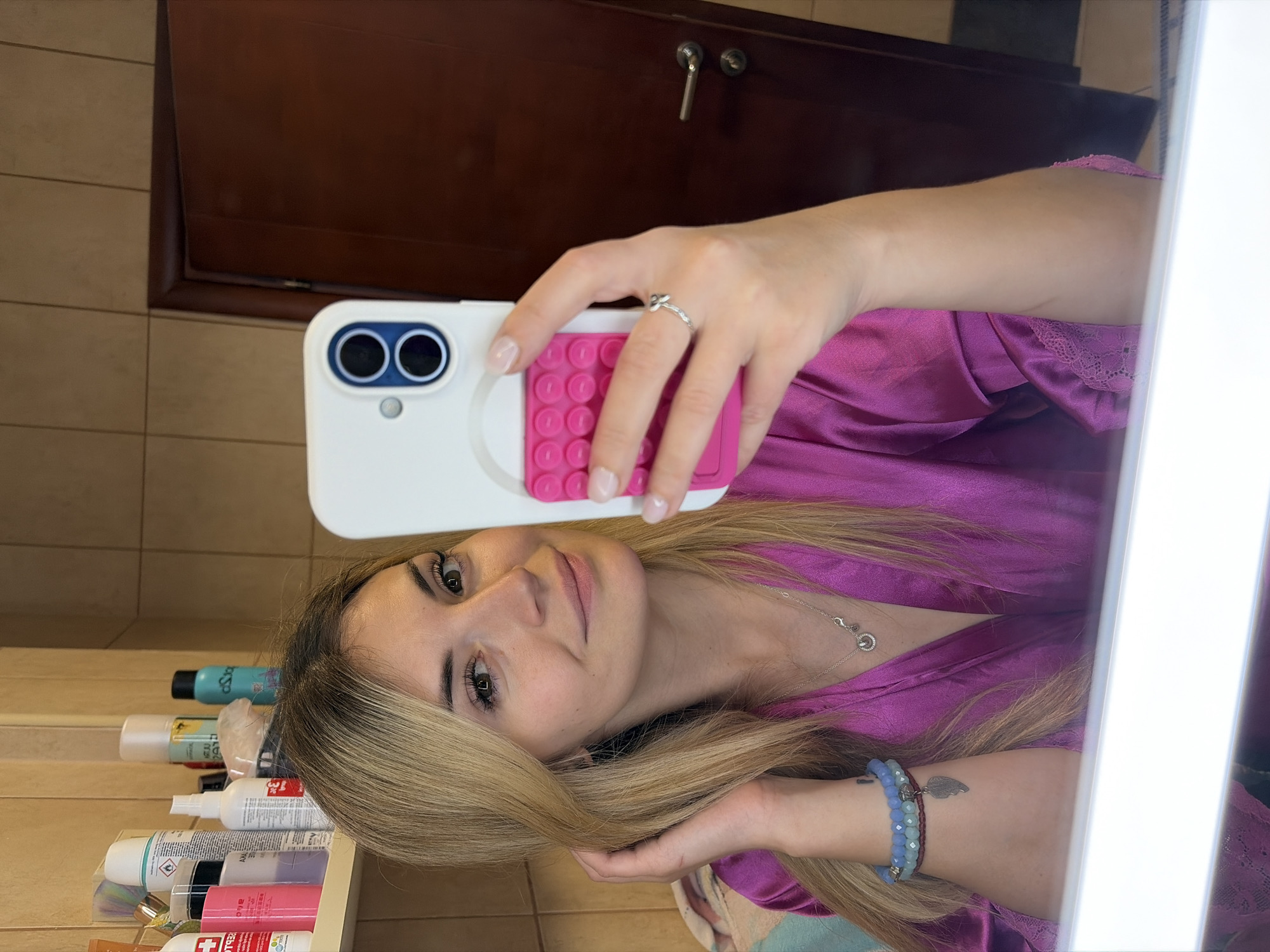 A woman in a magenta robe takes a selfie in what appears to be a bathroom. Her light blonde hair flows around her face, and she wears a silver necklace and blue beaded bracelets. She holds a smartphone with a white and pink case. Behind her, a shelf holds toiletries, and a dark wooden cabinet is visible on the wall. The location is Budva, Montenegro.