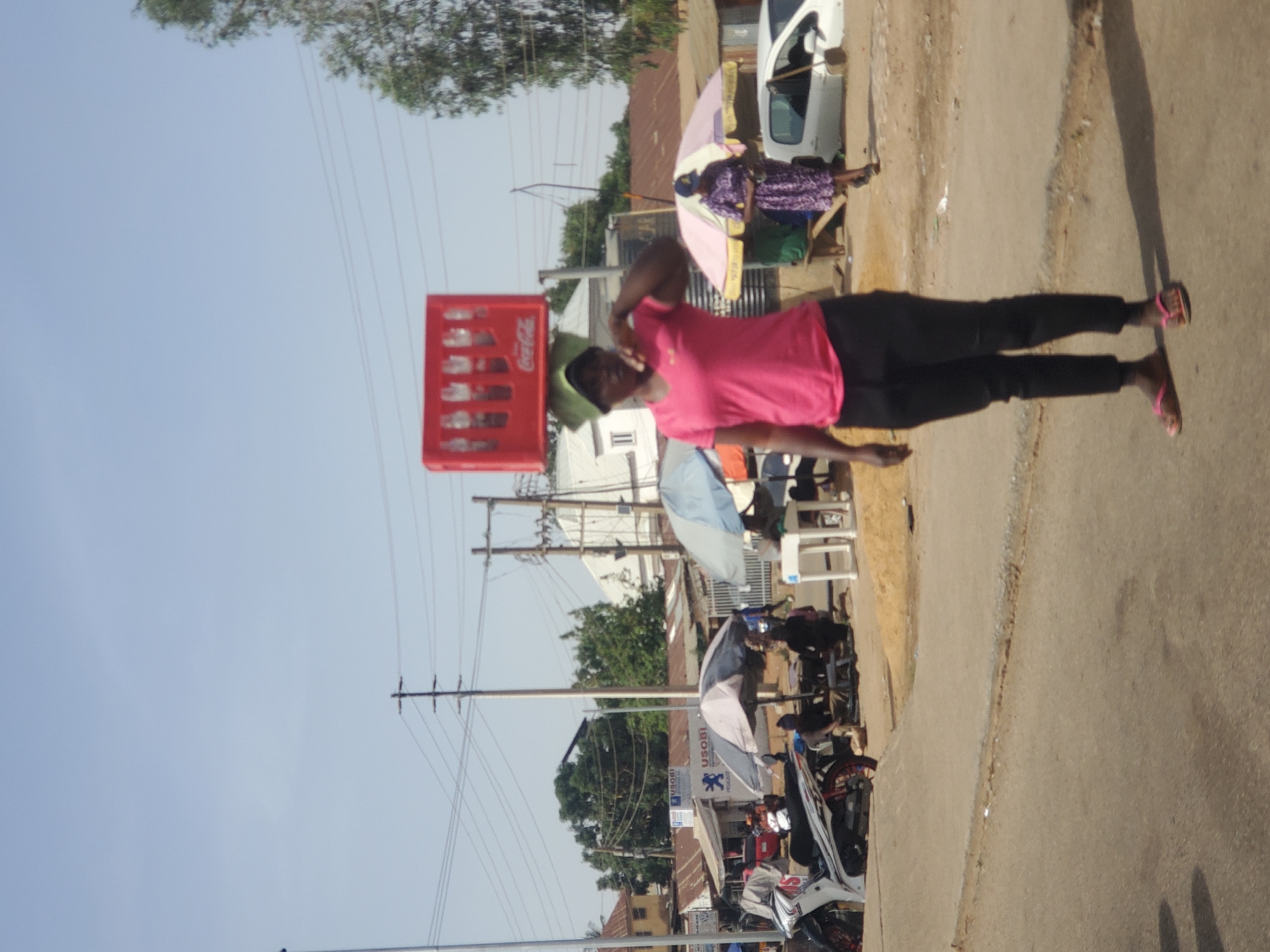 The image depicts a bustling street scene, likely in Jos, Nigeria, under a clear, bright sky, suggesting it is daytime with strong sunlight evident from cast shadows. The central focus is a person, wearing a pink t-shirt, dark trousers or leggings, and pink flip-flops, expertly balancing a red Coca-Cola crate on their head. Their right arm is slightly raised, possibly to steady the load. The Coca-Cola logo is clearly visible on the side of the crate.
In the mid-ground, to the right of the main subject, another individual is seated beneath a light purple and white umbrella, possibly attending a street stall. They are dressed in a patterned outfit. Further into the background, the scene reveals more street activity, including parked motorcycles and a white car.
The environment features a road surface that transitions from dirt to paved sections, flanked by buildings, many of which have corrugated iron roofs. Utility poles with numerous overhead wires are prominent against the sky. Another light blue and white umbrella is visible in the background, suggesting additional vendors or shaded areas. A sign on one of the buildings reads "USOBI" and includes what appears to be a Peugeot logo, indicating a local business. The overall image captures a slice of daily life, highlighting street vending and traditional methods of transport in a vibrant, sunlit urban setting.