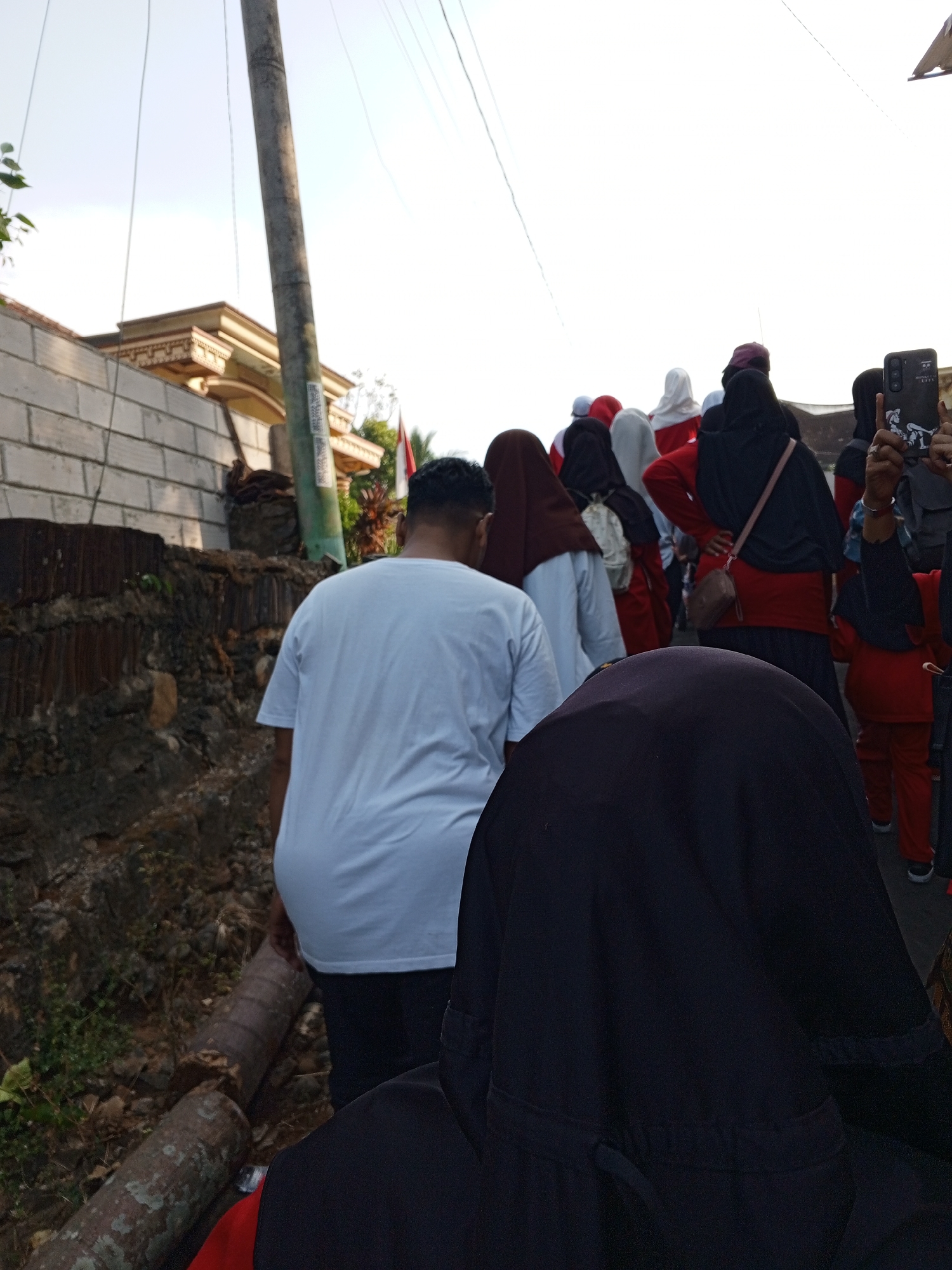 In Lemahbang, Indonesia, a group of people walk along a street. The foreground is dominated by the back of a person wearing a dark headscarf, obscuring their face. To their left, a person in a white t-shirt is partially visible from behind, facing away from the camera. Behind them, a line of individuals, mostly women wearing hijabs and headscarves in various colors like red, white, and brown, are walking down the street. Some are wearing red shirts and black skirts or pants. One person in the middle of the group is taking a photo with their phone. The street is lined with a low brick wall and some vegetation. A tall utility pole stands to the left of the frame. In the background, there's a glimpse of a building with decorative architectural features and what appears to be an Indonesian flag. The sky is bright and clear, suggesting it's daytime. The overall scene depicts a procession or a group of people moving through a residential area in Lemahbang.