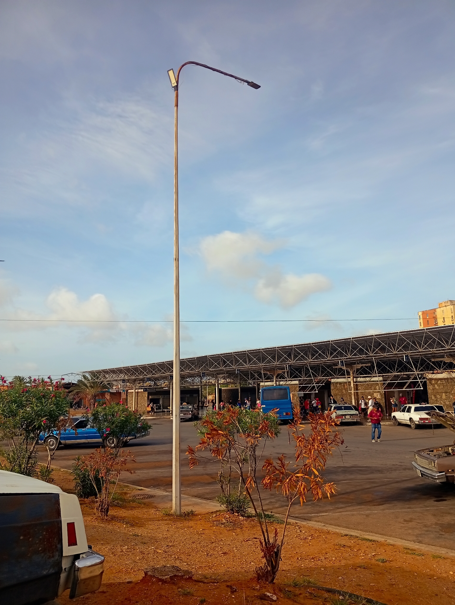 The image depicts a daytime scene at what appears to be a bus station or public transportation hub in Juan Griego, Venezuela. A tall, slender lamppost dominates the foreground, casting a shadow. Behind it, a covered waiting area with a metal truss roof structure spans across the frame, with several stalls or shops visible beneath. Multiple people are milling about in this area. A blue bus is parked to the left of the frame, and several cars, including a white sedan and a light-colored sedan, are also present in the vicinity. A person in a red shirt and blue jeans is walking through the open area. The ground is a reddish-brown, likely dirt or gravel, with some sparse vegetation, including bushes and small trees with dry, orange leaves. In the background, a multi-story building with orange and yellow coloring is visible. The sky is partly cloudy with a light blue hue. The overall impression is of a functional, somewhat weathered public space.