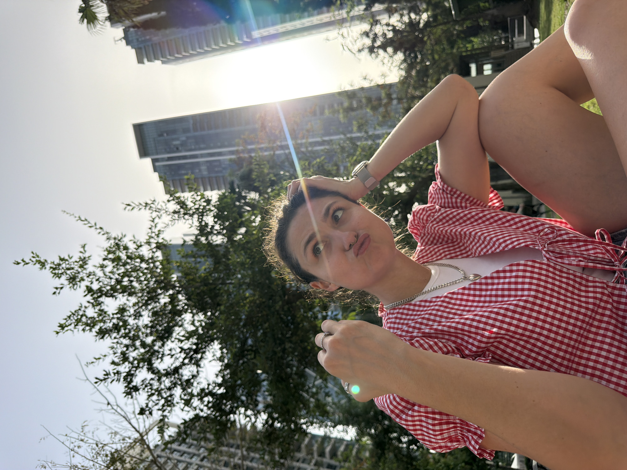 The image is an upward-tilted, medium shot of a young woman lying on the grass, seemingly in a park, with high-rise buildings visible in the background. The location is likely Tel Aviv, Israel, judging from the architecture. The woman has her legs bent and up, with one arm behind her head and the other extended, holding something small in her hand. She is wearing a red and white gingham patterned shirt over a white top, a silver chain necklace, and a watch on her left wrist. The background includes green foliage, trees, and modern, glass-faced skyscrapers. The sun is visible, creating a bright lens flare.