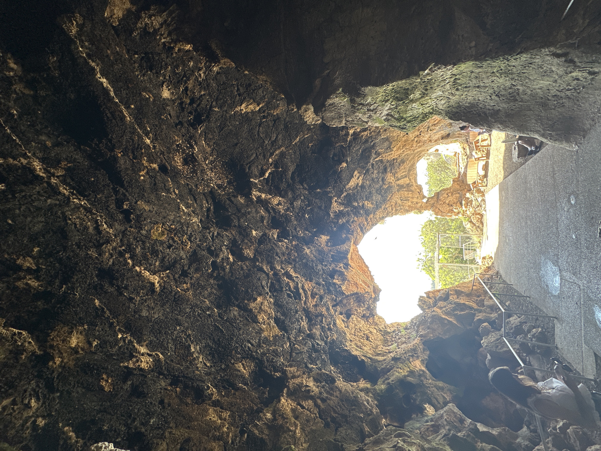The image depicts the interior of a cave, shot from below, looking upwards towards a hole that allows light to enter. The walls of the cave are textured with dark, rough rock, showing variations in color. A pathway and stairs are visible on the right side of the image, with people using them. The pathway appears to lead outside, indicated by brighter light and greenery. The location is potentially within the Hof HaCarmel Regional Council, Israel, based on the geographical context.