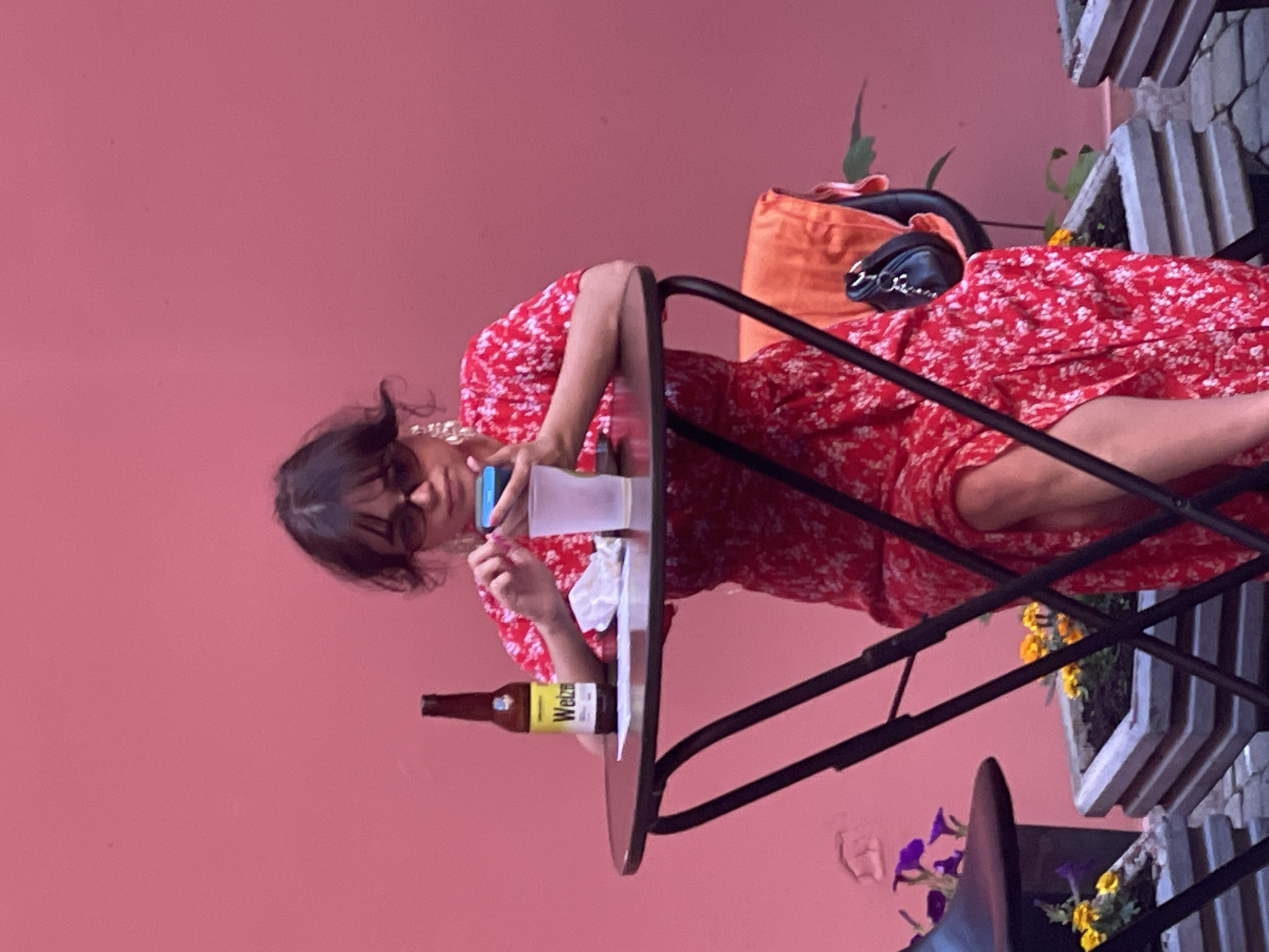 A young woman with dark hair, wearing sunglasses and a red floral dress, sits at a small round table with a black metal frame. She is holding a blue and white smartphone in her right hand and appears to be looking at it. Next to her on the table is a clear plastic cup and a crumpled white napkin. To the left of the cup is a brown beer bottle with a yellow label. The woman is seated in an outdoor setting with a vibrant pink wall as the backdrop. In the foreground, planters with yellow and purple flowers are visible, as well as the legs of the table and chairs. The overall scene suggests a casual, outdoor cafe or seating area in a city, potentially Saint Petersburg, Russia, given the query.