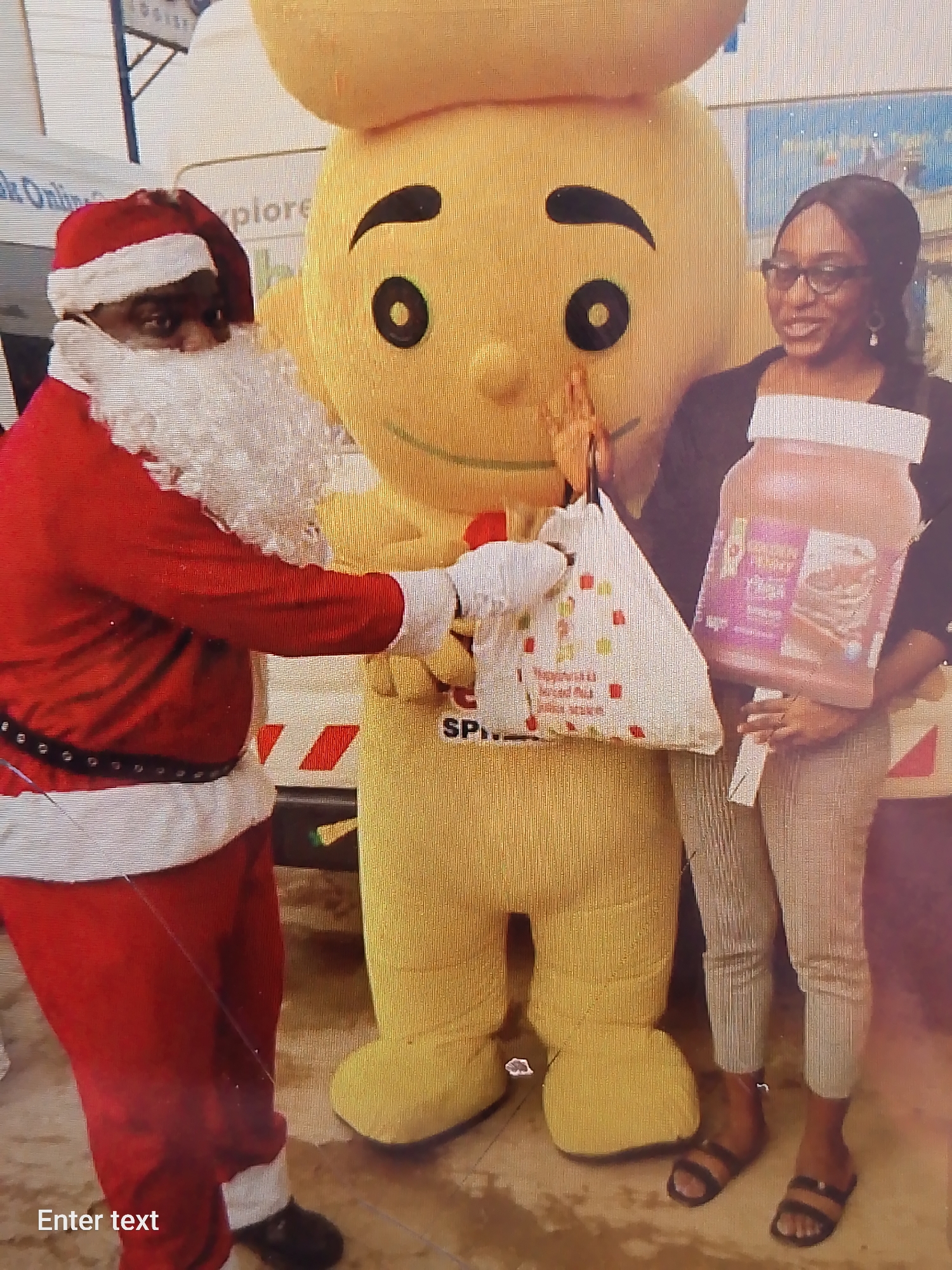 The image shows a scene in Adankolo, Nigeria. A person dressed as Santa Claus in a red suit with white trim hands a white shopping bag to a large yellow mascot. A woman stands to the right of the mascot, holding a large jar of what appears to be a food product. The floor looks like worn concrete. A vehicle, possibly a delivery truck, is visible behind the figures.