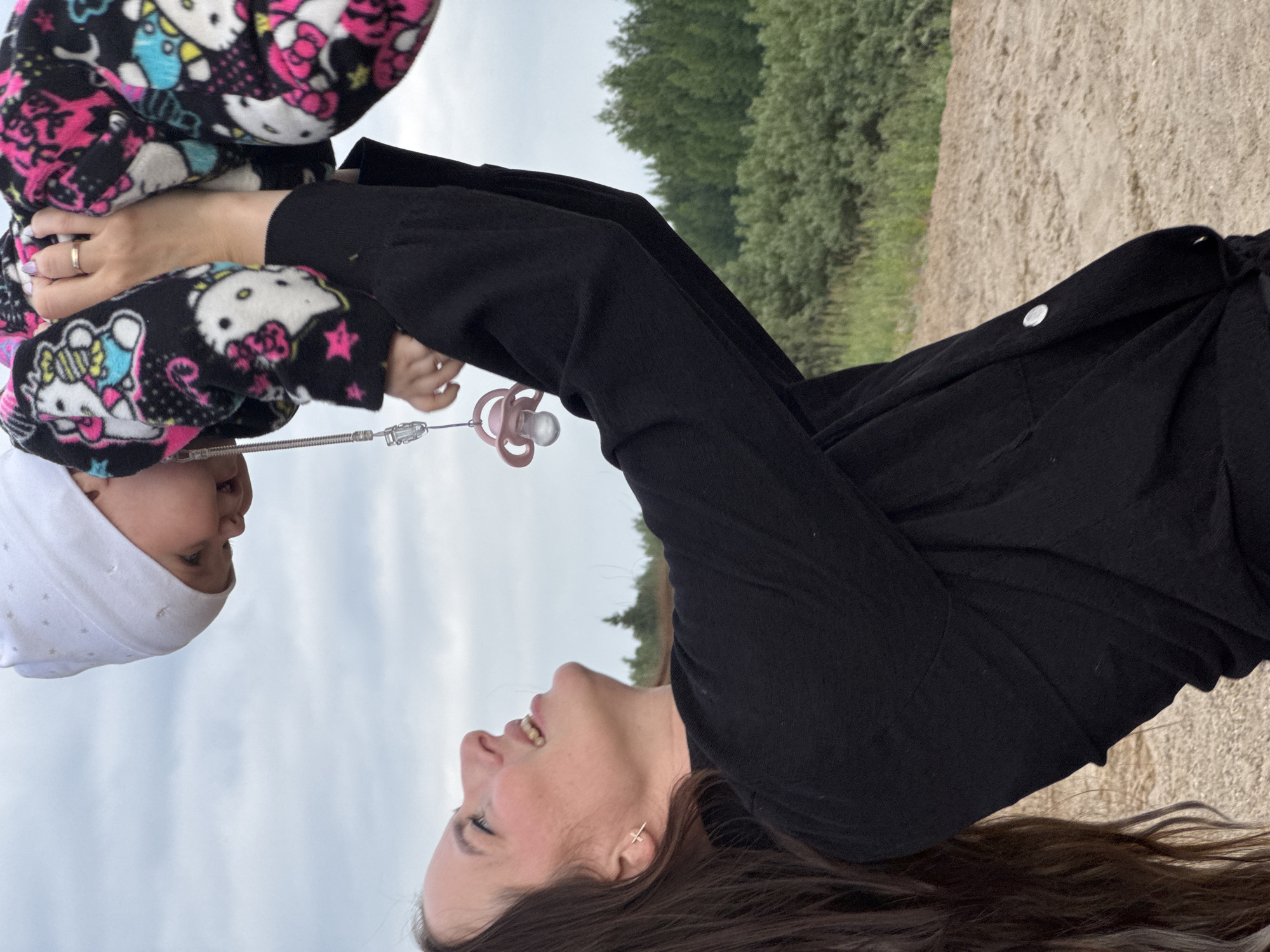 In Novyi, Russia, a young woman with long brown hair is holding a baby aloft in the air. The woman is wearing a black top and looking up at the baby with a smile. The baby is wearing a white hat and a black and pink patterned outfit featuring cartoon characters. A pink pacifier hangs from a string attached to the baby's clothing. They are outdoors, with a background of green trees and a cloudy sky. The ground beneath them appears to be a sandy or gravelly terrain.