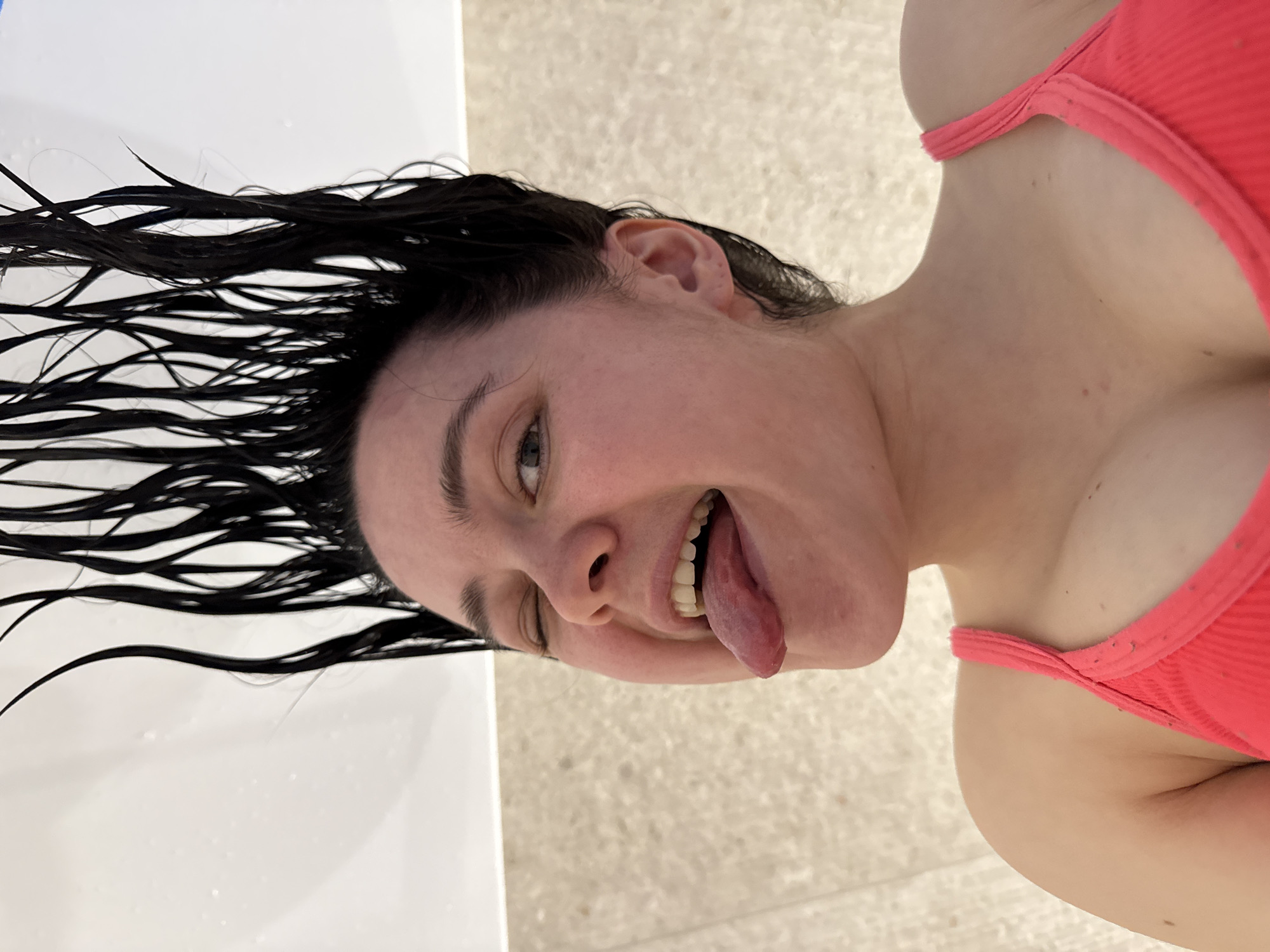 A close-up, tilted shot captures a person, likely a woman, in a playful moment. Her head is angled, and she looks upwards with one eye winking, her tongue playfully sticking out. She has wet, dark hair spread out on a white surface, possibly the edge of a bathtub or a pool. She is wearing a coral-colored, ribbed swimsuit top. The background features a light-colored, textured surface, resembling tiles, consistent with a poolside or bathroom setting. The overall mood is cheerful and carefree. This scene is likely taking place in Izhevsk, Russia. There is no visible text in the image.