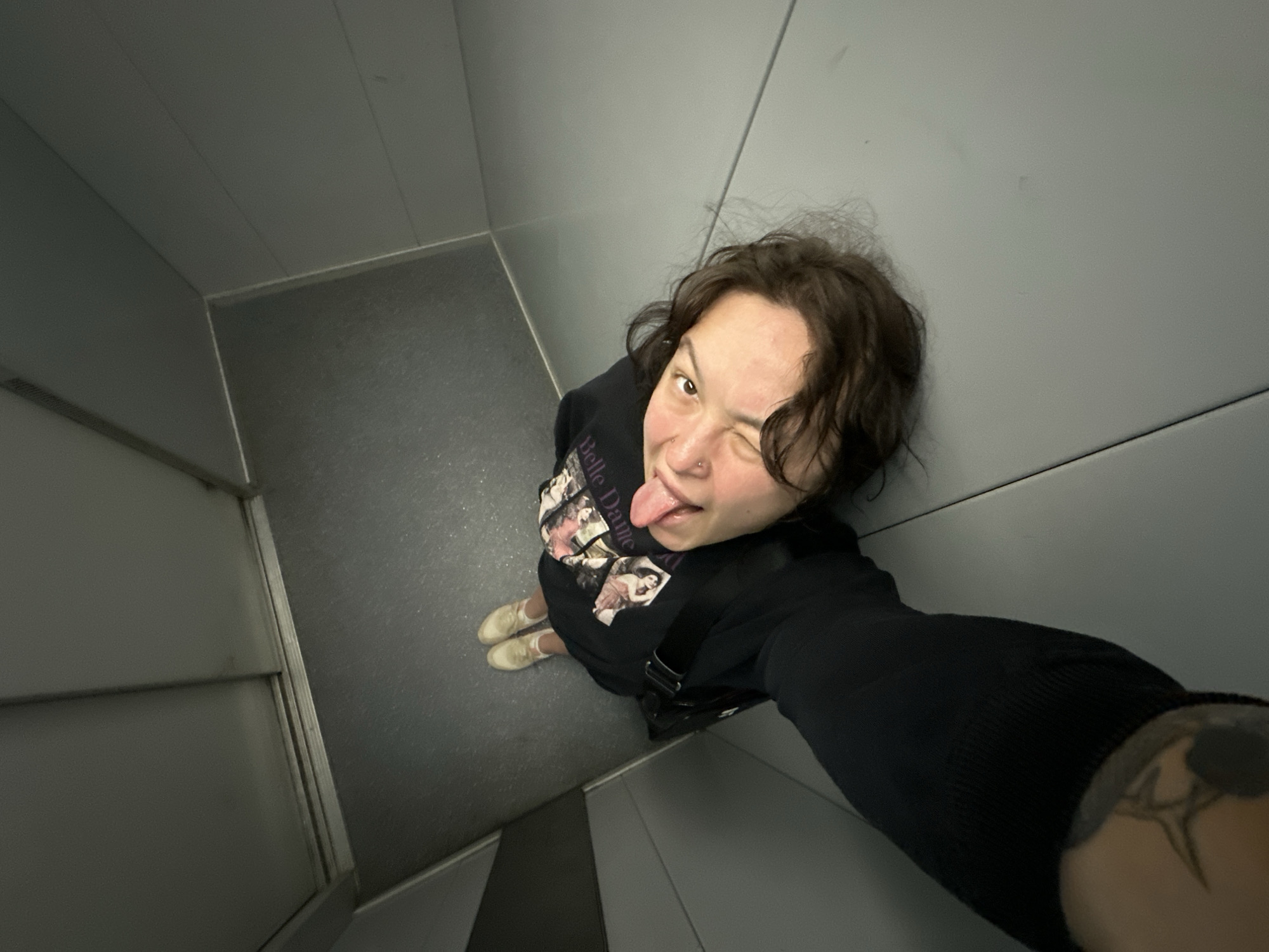 Within an elevator in Chaikovsky, Russia, a low-angle selfie captures an individual looking directly at the camera with a playful expression, tongue extended, and their left eye partially closed in a wink. The person has medium-length dark brown hair, fair skin, and a visible septum piercing. They are wearing a dark long-sleeved top adorned with multiple graphic prints of vintage-style female portraits and light-colored, off-white lace-up sneakers. A dark shoulder bag is positioned next to their left hip. The arm holding the camera, partially visible in the foreground, displays an abstract dark ink tattoo on the inner forearm. The elevator interior features light gray paneled walls with visible vertical seams and a darker gray floor with distinct seams and minor speckling. The upper left portion of the frame shows the top edge and track of a closed elevator door.