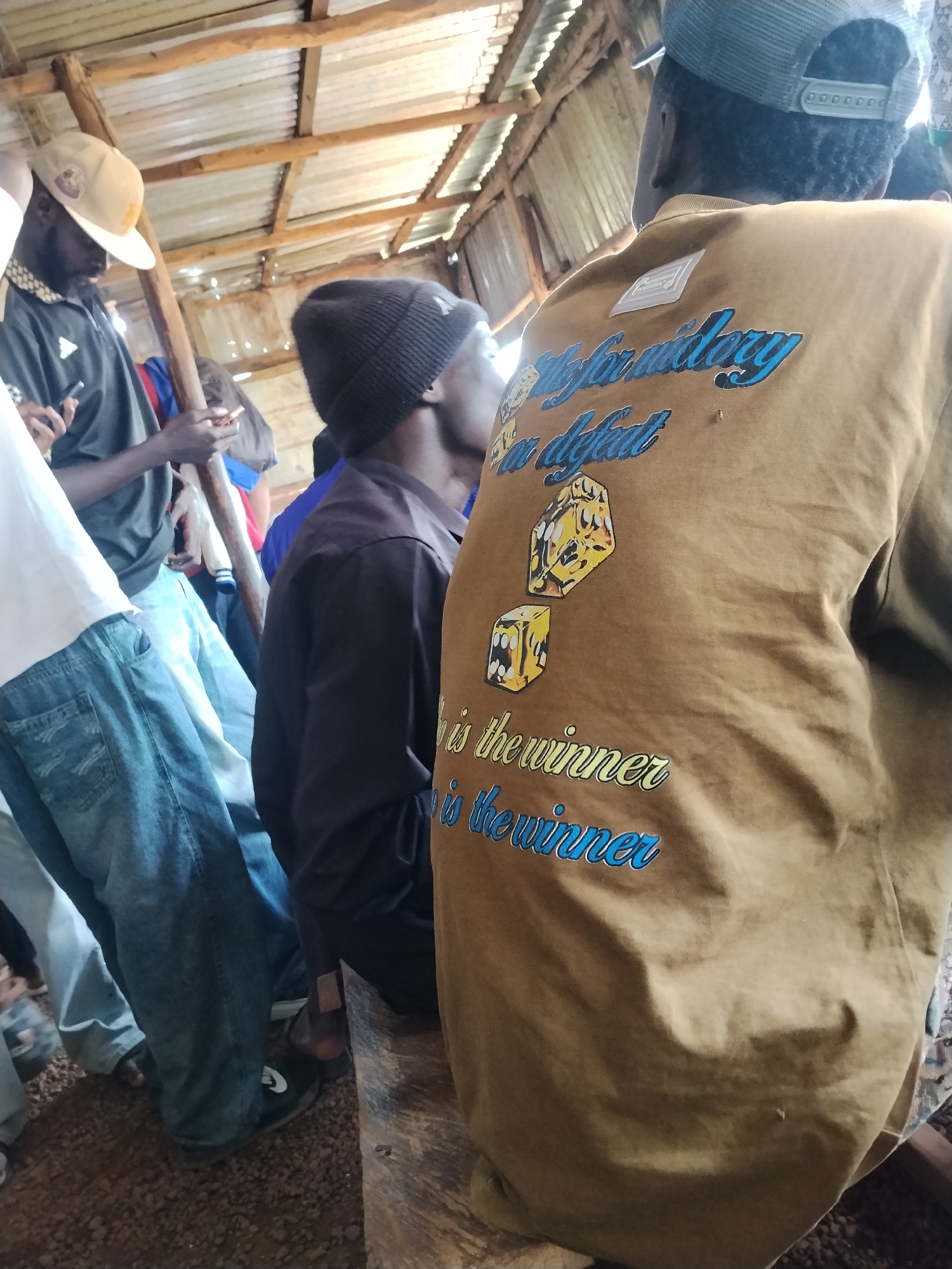 The image depicts a group of individuals gathered inside a rustic wooden structure, possibly a shelter or market stall, with a corrugated metal roof supported by wooden beams. The environment appears to be outdoors, under a covered area. The ground is covered with gravel and dirt.
In the foreground, a person wearing a tan t-shirt with blue and yellow text and graphics is prominently featured from the back. The text on the t-shirt reads "for victory" and "is the winner the winner". There are graphic depictions of dice on the t-shirt.
To the left, another person is seen wearing a dark t-shirt and a beige cap. They are holding a mobile phone in their right hand and a cigarette in their left. Further to the left, a portion of a person wearing a white t-shirt and blue jeans is visible.
Another individual is wearing a black beanie and a dark long-sleeved shirt. The people are standing and facing away from the camera in various directions. The overall atmosphere seems casual and informal. The location is specified as Durumi, Nigeria.