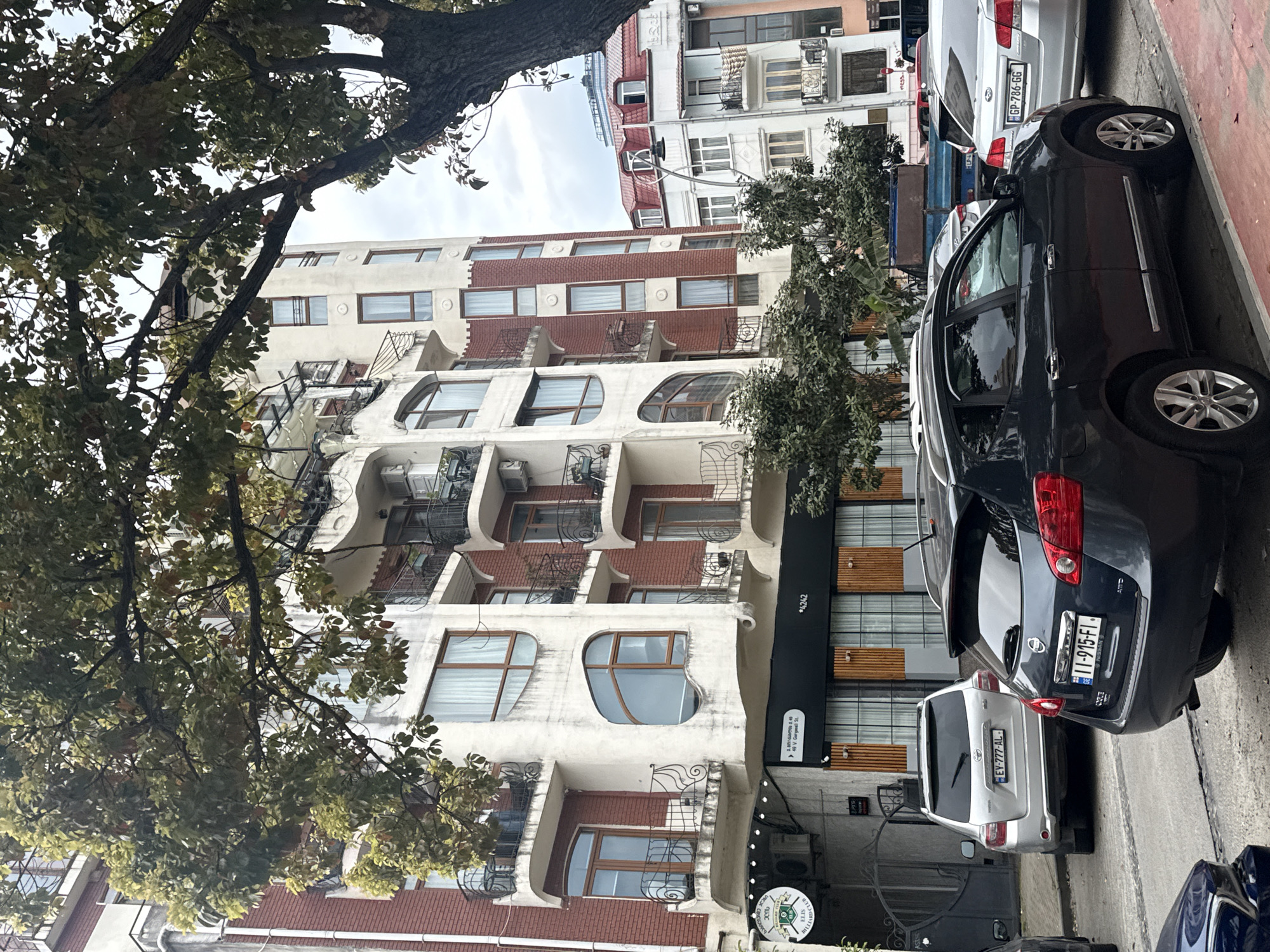 The image captures a street scene in Batumi, Georgia, showcasing distinctive architecture, parked cars, and lush greenery under an overcast sky. The photograph is rotated 90 degrees clockwise, with the street and vehicles running horizontally across the lower part of the frame, and buildings extending vertically.
The prominent structure in the center is a multi-story building characterized by an unusual, organic architectural style. Its facade features undulating white concrete sections interspersed with red brickwork. Numerous balconies with decorative black wrought-iron railings and several air conditioning units are visible. Many windows are uniquely shaped, featuring rounded or elongated curved designs, contributing to the building's distinctive appearance.
A large tree with dense green foliage partially covers the upper left portion of the building, its branches extending across the frame. Smaller trees and shrubs are also visible along the street.
Along the curb, several cars are parked. A dark grey Nissan SUV is prominent, displaying the Georgian license plate "11-915-FI" and the text "AWD". Behind it is a silver SUV or sedan with license plate "EV-777-AL-". Further back, another car shows the plate "GP-786-GG".
At the ground level of the main building, commercial signage is visible. A round sign reads "BILLIARD CLUB" and "ELIS". Another black sign, partially obscured, shows "49 V. Gergasali St." and the number "4242". The sky above is overcast, indicating a cloudy daytime setting. No people are visible, but the presence of parked cars and businesses suggests an active urban environment.