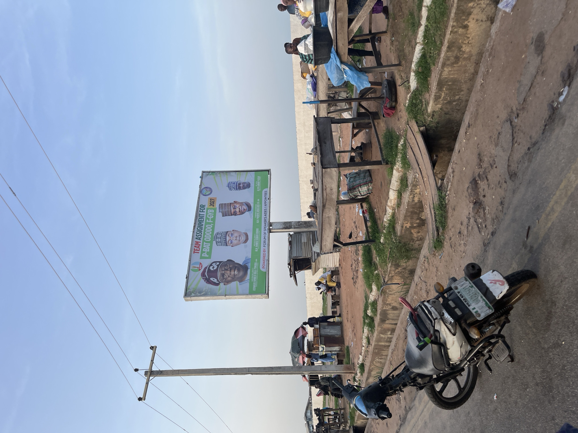 This media file depicts an outdoor scene with a billboard in the foreground, likely promoting a political campaign for the year 2027, featuring candidates' portraits and the text "TEAM ASSIGNMENT FOR P-BAT ODODO, F-GYB 2027". The billboard also includes names like "ASIWAJU AHMED BOLA TINUBU" and "ALIYU UMAR YUSUF". A motorcycle is parked on the right side of the image, with its license plate partially visible. In the background, several people are gathered, some sitting and others standing. The setting appears to be a street or roadside area with some makeshift structures and a drainage ditch. Power lines and a utility pole are also visible in the sky. The overall lighting suggests it might be late afternoon or early morning. The scene seems to be in a somewhat arid environment, with dusty ground and sparse vegetation. There are no clear indicators of the specific location.