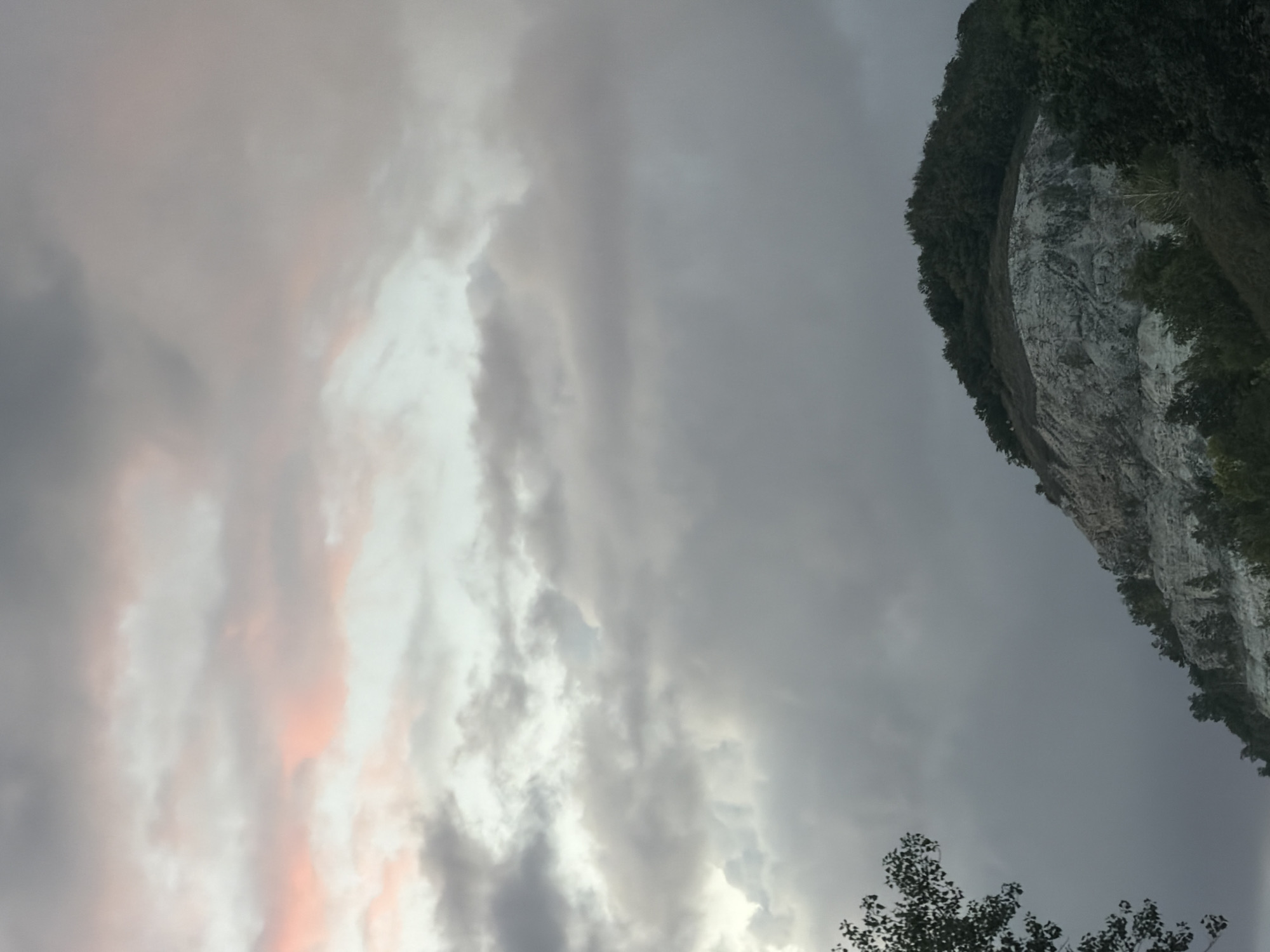 The image depicts a view of a rocky hill covered in vegetation, with a sky filled with clouds. The hill is characterized by its rough, stony texture and is dotted with trees and bushes. The clouds in the sky vary in color from light grey to pinkish hues, suggesting it might be sunrise or sunset. In the foreground, on the left side of the image, there is a silhouette of a tree. The overall impression is a natural landscape scene, likely taken outdoors. The location is identified as being in the city of Samara, Russia, though no specific landmarks are identifiable within the frame. There are no people or discernible actions taking place in the image.