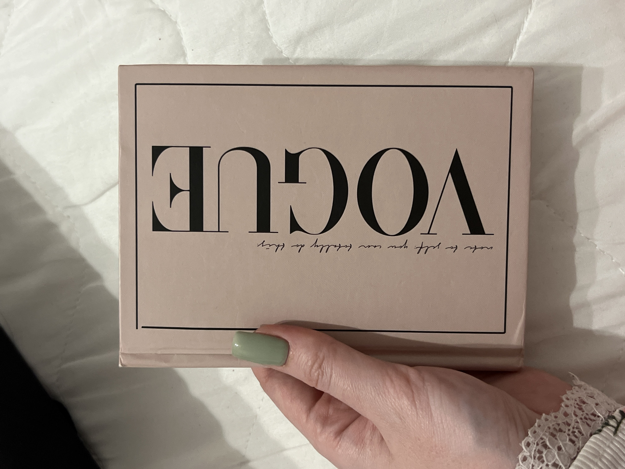 A hand with a light green painted nail holds a pale pink book with black lettering. The text "VOGUE" is printed vertically in large black letters on the right side of the book. Below the "VOGUE" text, in smaller cursive black letters, it reads "note to self, you can totally do this." The book is closed and positioned at an angle, with its spine facing left. The background is a white quilted surface, possibly a bedspread or blanket. The image was taken indoors. The location in Kazan, Russia cannot be determined from the provided image.
