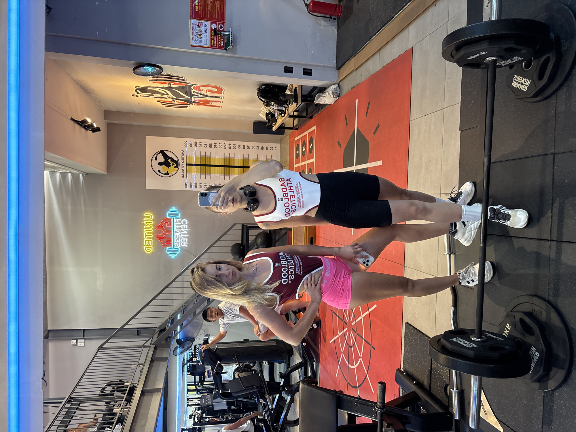 Here is a detailed description of the image:
Two young women are posing for a selfie in a gym setting. The woman closer to the camera has blonde hair, wearing a dark red tank top with white trim and a phrase that includes the word “BADBLOOD,” and pink athletic shorts. She is holding her phone out, presumably taking the photo. The other woman, behind her, has shorter blonde hair and is wearing a white tank top with similar dark trim and lettering with black athletic shorts. She is holding her phone and looking at the camera as well.
In the background, a man is working out on a treadmill. Gym equipment is visible throughout the scene. A staircase with metal railings is also present in the lower left corner of the image. Gym related signage and graphics are displayed on the walls, including a height chart and the logo for "UNITED FITNESS CENTER." The image also contains a barbell with black weight plates. The setting appears to be a commercial gym.
The text in the image, including the gym name and details on the clothing, may indicate that the image was taken in Đà Nẵng, Vietnam, as some of the text is in Vietnamese.