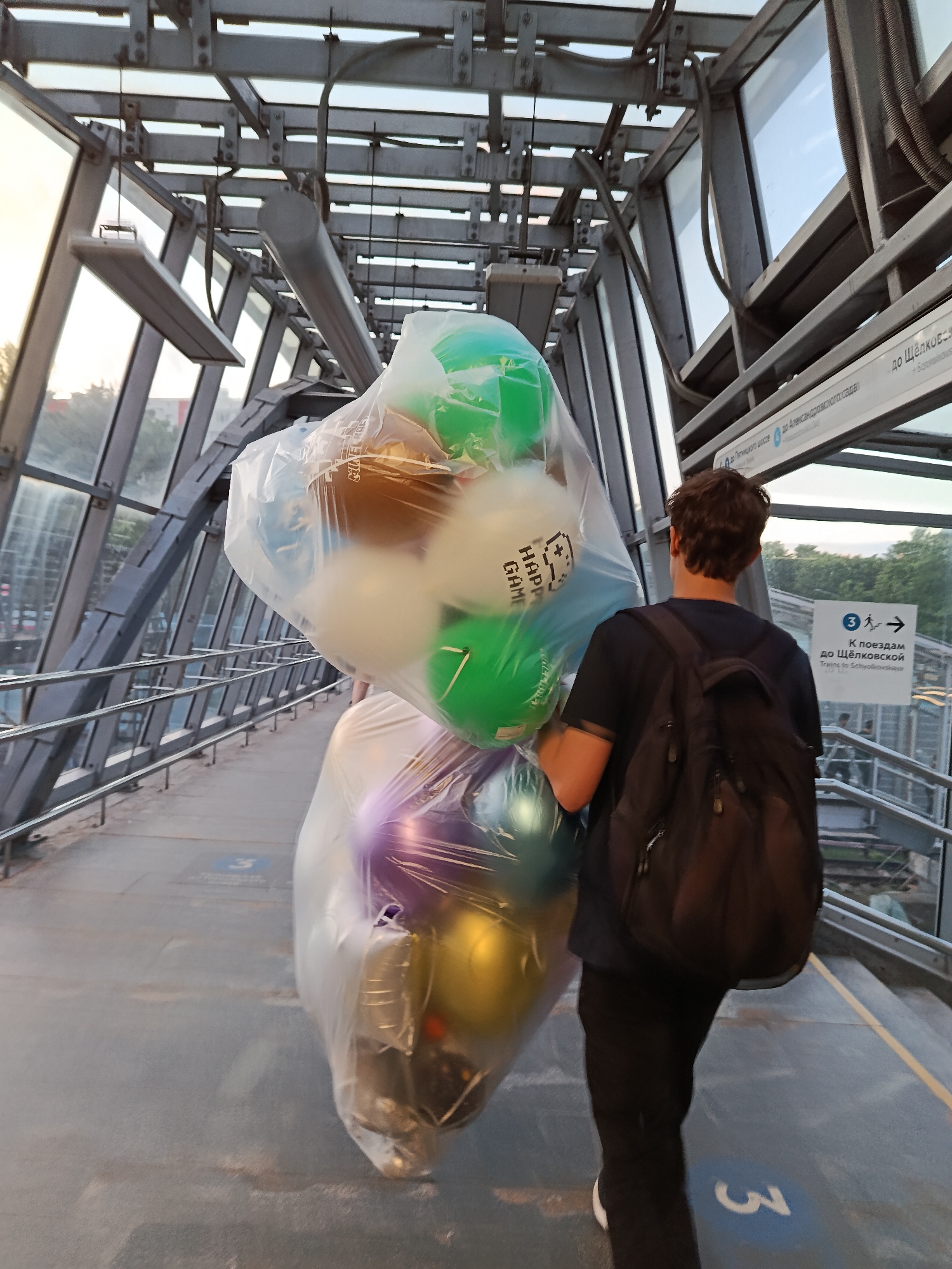 A person, viewed from behind, walks through a modern, enclosed pedestrian walkway or bridge, carrying a large, buoyant bundle of balloons. The individual wears a dark t-shirt, black pants, and a black backpack. The extensive collection of balloons, contained within clear plastic bags, is held in front of the person, partially obscuring their body. The balloons display a variety of colors, including green, white, blue, brown, gold, and purple, with some appearing metallic and others opaque. Text visible on one white balloon reads "HAPPY GAME".
The setting is a pedestrian overpass or skywalk, characterized by a robust metallic framework and large glass panels that offer views of the outside. Distant trees and buildings are visible through the windows, under what appears to be a bright, possibly overcast sky. The walkway's floor is a light gray, with a blue circle marking containing the number "3" and an arrow visible on the ground.
Signage is present on the right side of the walkway. One sign, in both Russian and English, directs "К поездам до Щёлковской" / "Trains to Schyolkovskaye," indicating a direction to a specific train station or line. Another sign, partially visible further along the wall, mentions "до Пятницкого шоссе до Александровского сада" (to Pyatnitskoye Highway to Alexandrovsky Garden), suggesting a location within Moscow, Russia. The time of day appears to be daytime, with ample natural light entering the structure.