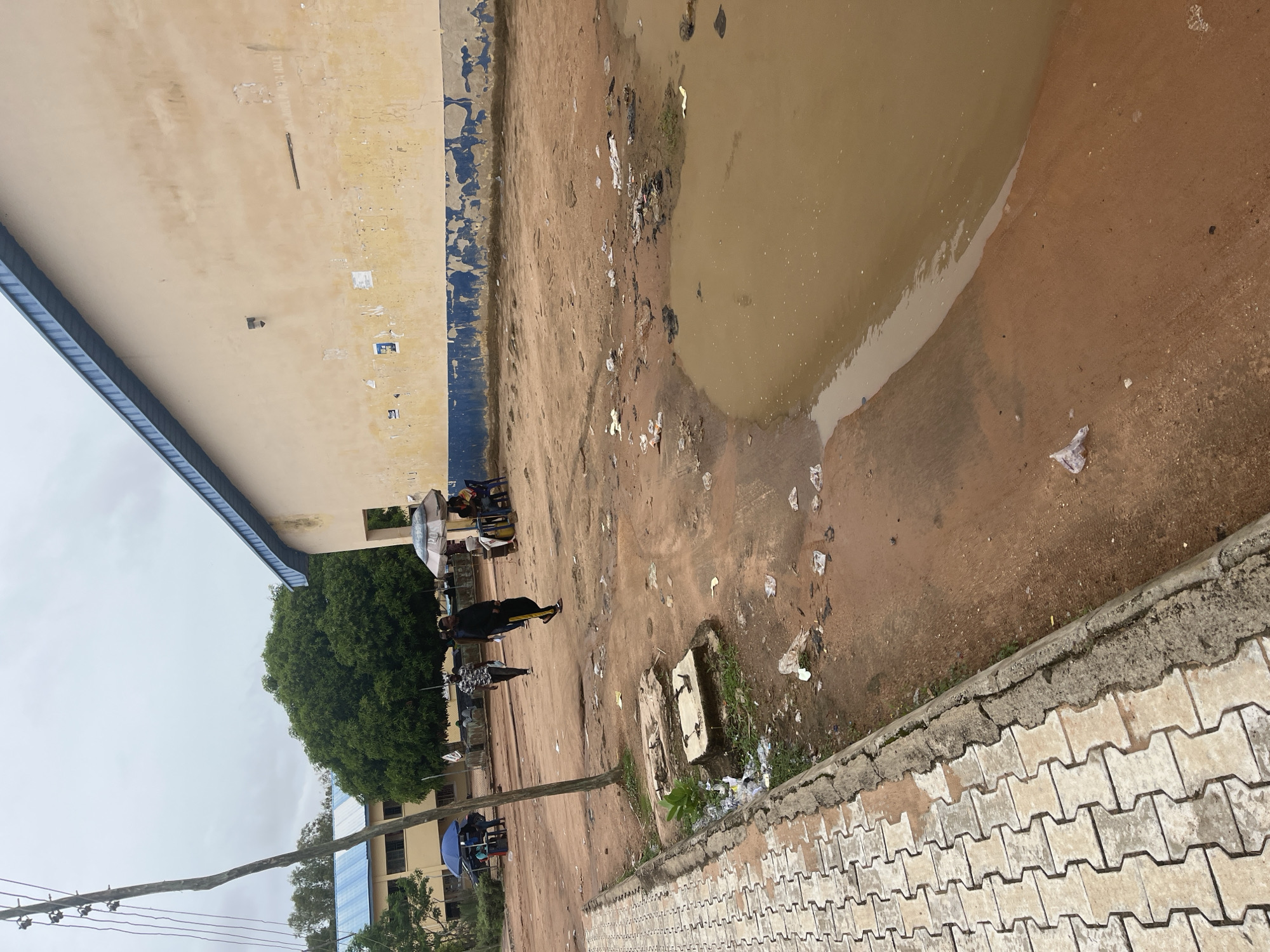 The image depicts an outdoor scene, likely on the grounds of a school or public institution in Jalingo, Nigeria. The ground is primarily a reddish-brown dirt, with a large, muddy puddle occupying the right side of the frame. There is evidence of litter scattered across the ground, including plastic bags and other debris. A paved walkway, made of interlocking concrete blocks, runs along the left edge of the image, bordering the dirt area.
In the background, there are two school-like buildings with light-colored walls and blue-pitched roofs. Several trees, including a large, leafy one, are visible between the buildings and in the distance. A utility pole with wires stands on the left side of the image.
Several people are present in the scene. Some are walking across the dirt area, while others are seated under umbrellas, possibly at makeshift stalls. One individual in the foreground is walking away from the camera, wearing dark clothing with yellow stripes on the pants. Another person is walking towards the camera in a patterned shirt and dark pants. Near the building on the right, a group of people are gathered around tables and chairs, some of whom are under a white umbrella. A blue umbrella is also visible near the building on the left. The sky is overcast, suggesting a cloudy day.