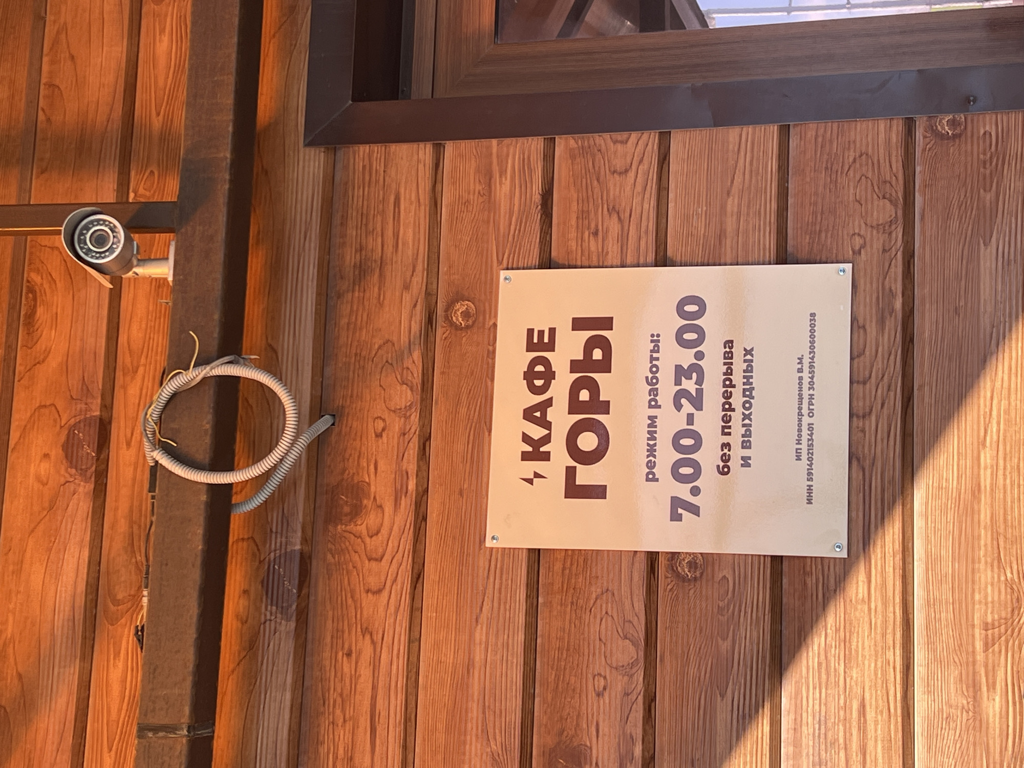 The image shows a close-up of a wooden exterior wall with a sign in Russian for "Cafe Gory" (meaning "Cafe Mountains"). The sign states the cafe's opening hours as 7:00 - 23:00 and indicates that it is open every day without breaks. The sign also includes the business registration information. A security camera is mounted on a post to the left of the sign with some exposed wiring. The location is Gory, Russia.