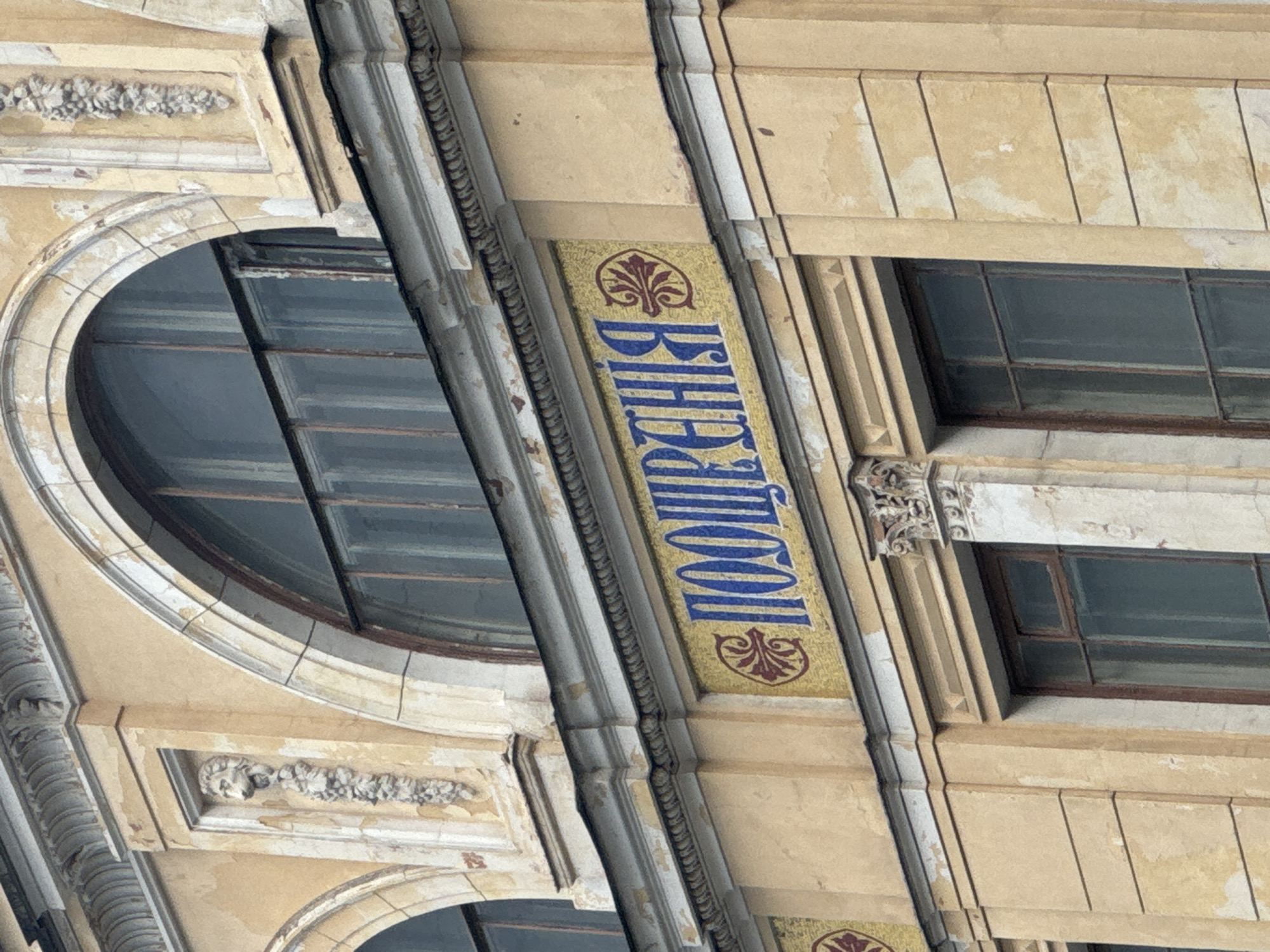 The image shows a detail of the facade of a historic building in Saint Petersburg, Russia. The building features decorative architectural elements and is adorned with a mosaic inscription. The inscription, rendered in blue Cyrillic letters on a yellow tiled background, reads "ПООЩРЕНИЯ" which translates to "Encouragement". The mosaic is bordered by decorative motifs in red and yellow. Above the inscription, there are arched windows with ornate frames and decorative carvings. Below the inscription, there are windows with darker frames, and the building's facade is painted in a pale yellow color, showing signs of age and wear. The overall impression is one of grand, historical architecture.