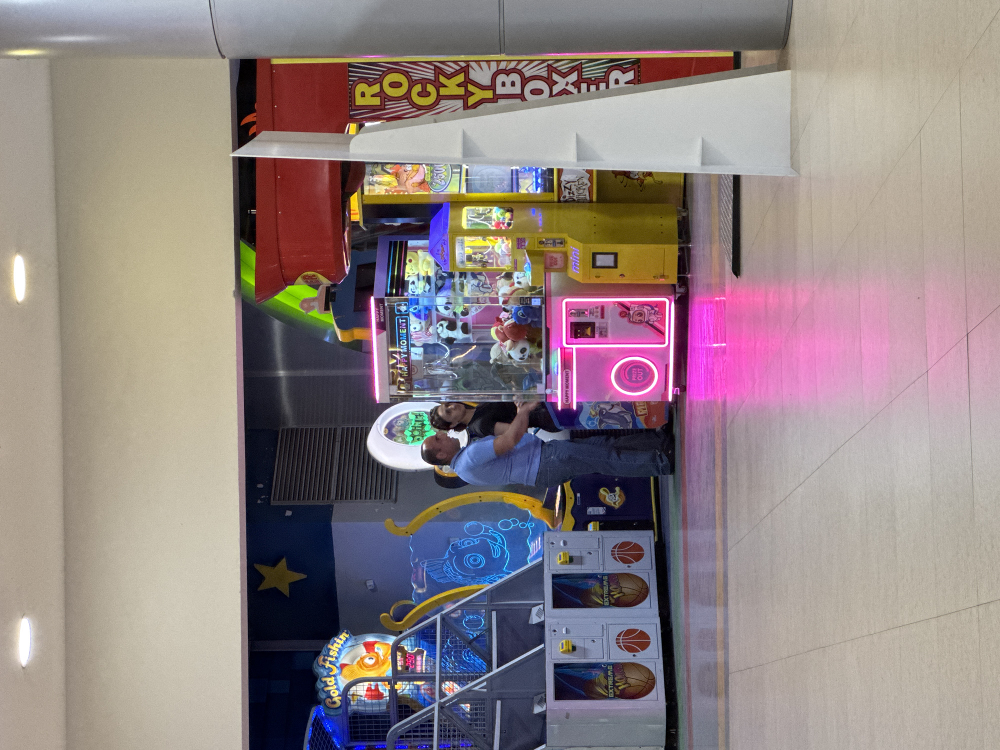 An indoor arcade or entertainment center is brightly lit by the colorful glow of various gaming machines. The setting features a light-colored tiled floor and neutral walls, with a dark blue section decorated with a yellow star.
Two adult men are visible in the midground, engaged with the arcade games. One man, wearing a light blue polo shirt and jeans, stands with his back partially to the viewer, looking towards a pink-lit claw machine. Another man, in a dark t-shirt, is positioned closer to the same machine, his face partially obscured, also appearing focused on the game.
The arcade is filled with a variety of machines. Prominently featured is a red and yellow "ROCKY BOXER" punching game. Nearby are several claw machines, including a yellow one labeled "mini" and another, pink-lit machine with plush toys inside, displaying "HAPPY MOMENT" and "PRIZE OUT" signs. Another game, partially visible, has text that appears to read "Dizzy Chicken". To the left, a "Gold Fishin'" game with a large fish motif is seen, along with multiple "EXTREME HOOP" basketball shooting games. The pink neon light from one of the machines casts a vibrant glow across the floor.