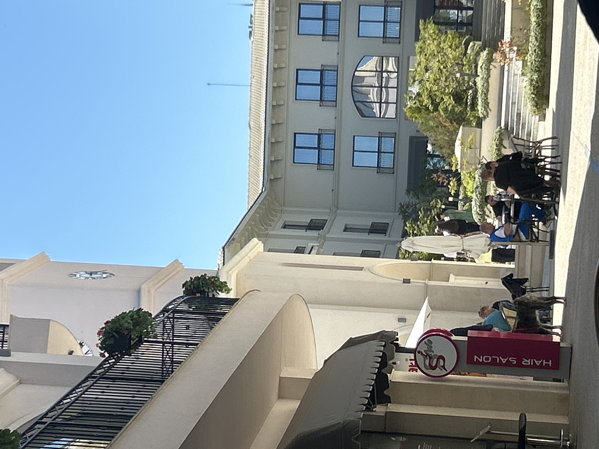 Here is a detailed description of the image:
The image captures an outdoor scene in an urban environment, likely in a city with a Mediterranean or European aesthetic. The sky is clear and bright blue. A multi-story building with classical architecture is visible, featuring multiple windows with dark frames and decorative elements on the façade. A lower, white building with an arched structure is visible to the left, adorned with a wrought-iron staircase and flower pots.
In the foreground, there is a sign for a "HAIR SALON" with white text on a red background, accompanied by a round logo. People are seated at tables under parasols near the building, suggesting a café or outdoor dining area. The people appear to be relaxed, engaging in activities like eating or conversing. There are chairs and tables positioned in the outdoor space, creating a casual and inviting atmosphere.