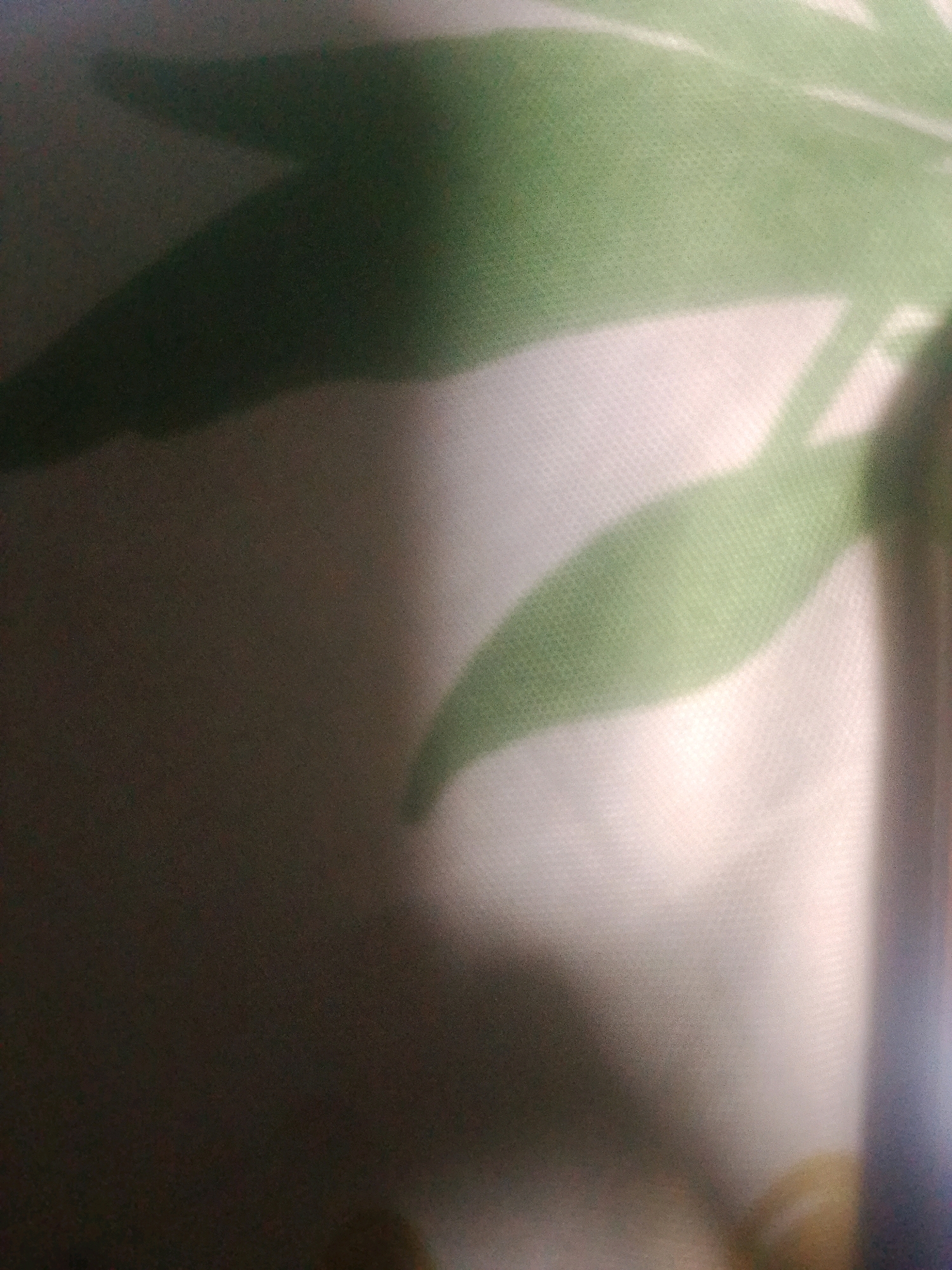 The image shows a blurred, close-up view of a patterned fabric, possibly a curtain or upholstery. The fabric has a light-colored background with a subtle texture, and overlaid on this are abstract shapes in various shades of green, suggesting a leaf or floral pattern. There is also a significant area of shadow, casting a dark, amorphous shape across the fabric. The lighting is uneven, with some areas brighter than others, creating highlights and deep shadows. The focus is soft, with no sharp details visible. The scene appears to be indoors, given the presence of fabric and indoor lighting. Without further context or clear landmarks, it is not possible to determine the specific location within Kecamatan Haurgeulis, Indonesia.