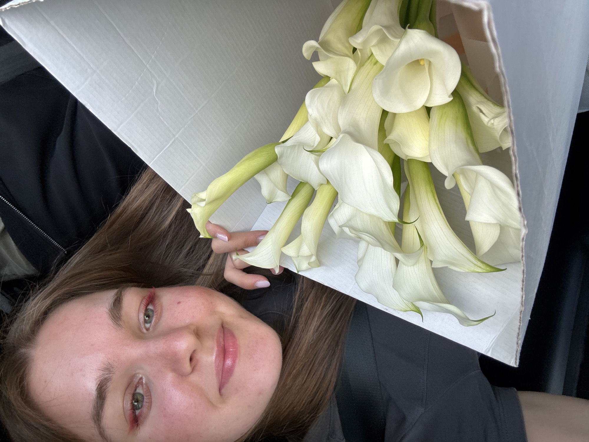 In the city of Perm, Russia, a young woman with long brown hair and green eyes smiles as she holds a bouquet of white calla lilies. She is wearing a dark grey t-shirt and a black jacket. Her makeup includes pink lipstick and red eyeshadow. The bouquet is presented in a white cardboard box. The background appears to be the interior of a vehicle, suggested by the seatbelt visible on the left side of the frame and the dark upholstery behind the woman.
