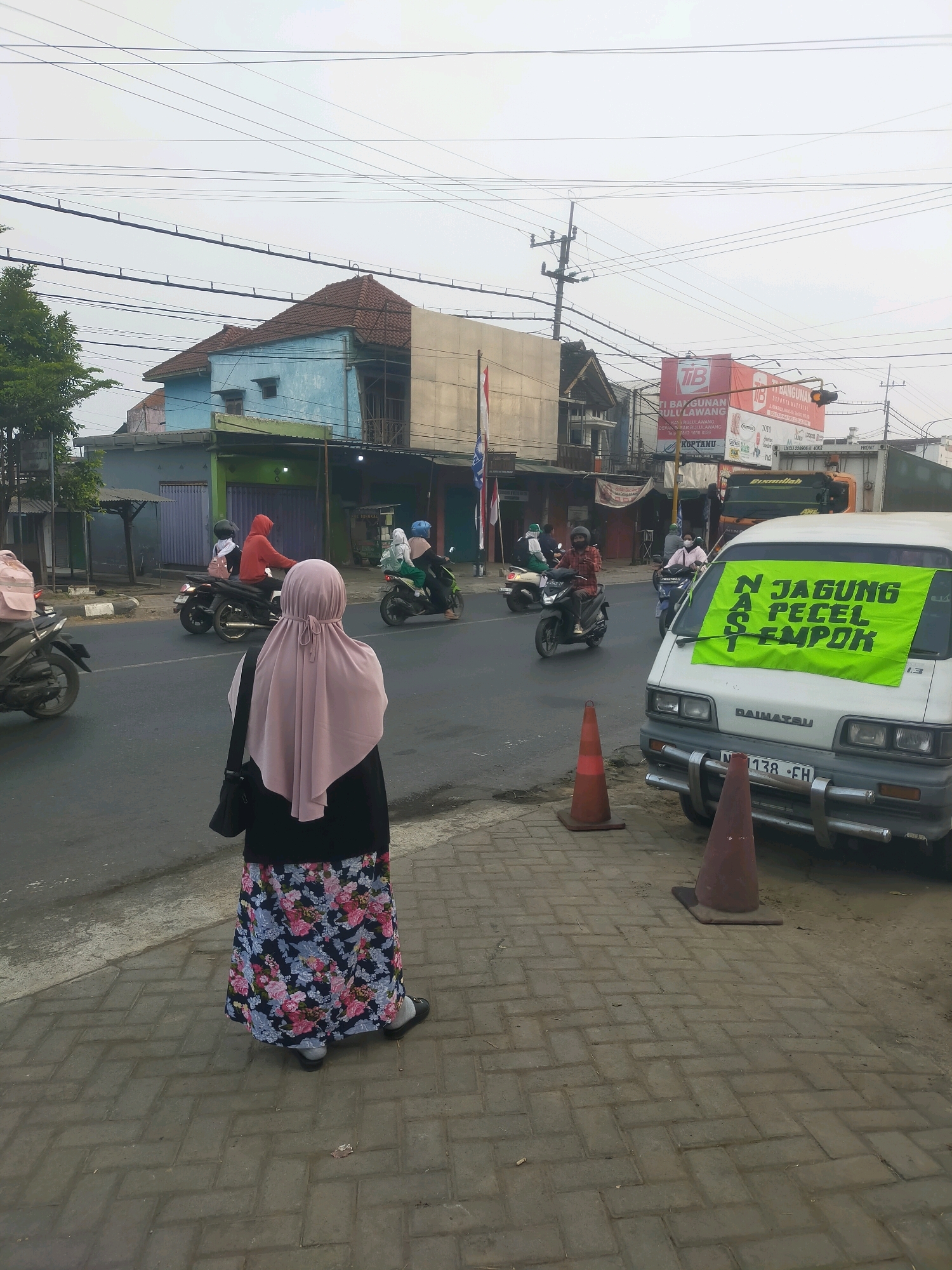 This is a medium shot of a street scene in Bululawang, Indonesia, taken from a slightly elevated perspective. The sky is overcast, and the scene appears to be during daylight hours. In the foreground, a woman wearing a pink hijab and a long, floral patterned skirt stands facing away from the camera, looking towards the street. She has a dark bag slung over her shoulder. She is standing on a paved sidewalk made of interlocking bricks.
The street in front of her is asphalt and has several motorcycles with riders. One motorcycle in the middle ground has a rider in a red hoodie. To the right of the street, a white van is parked. A bright green banner is attached to the windshield of the van, with bold black text that reads "NAS JANGUNG A PECEL TEMPOK". Two orange traffic cones are placed on the sidewalk in front of the van. In the background, there are buildings lining the street, including a two-story building painted light blue, and another building with a red and white sign that says "TB BANGUNAN". Power lines are visible overhead, crisscrossing the sky.