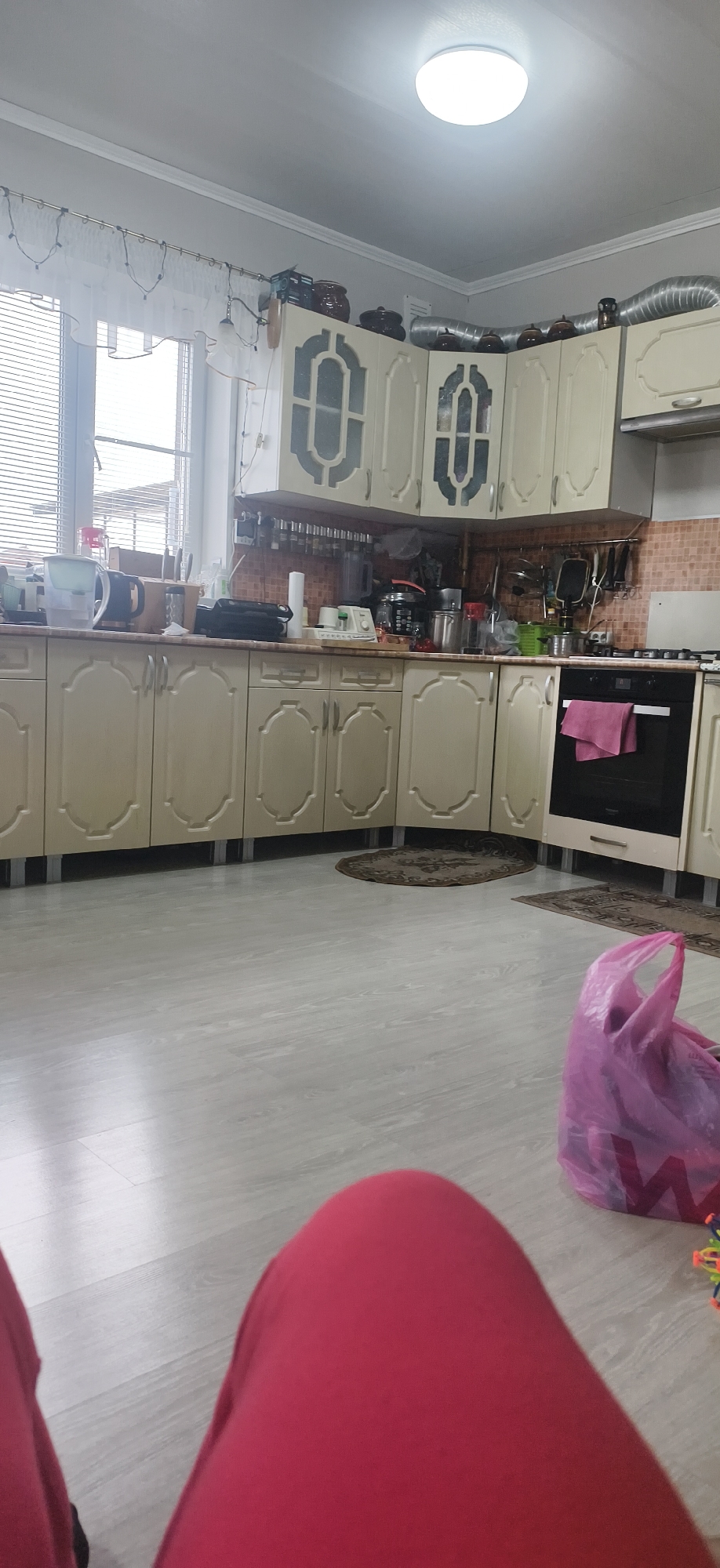 The image shows a kitchen with light-colored cabinets and appliances. There is a window with blinds to the left. The floor is light-colored and there is a rug near the cabinets. A pink bag is visible on the floor, along with a glimpse of someone wearing red clothing. The location is Фоки, Russia.