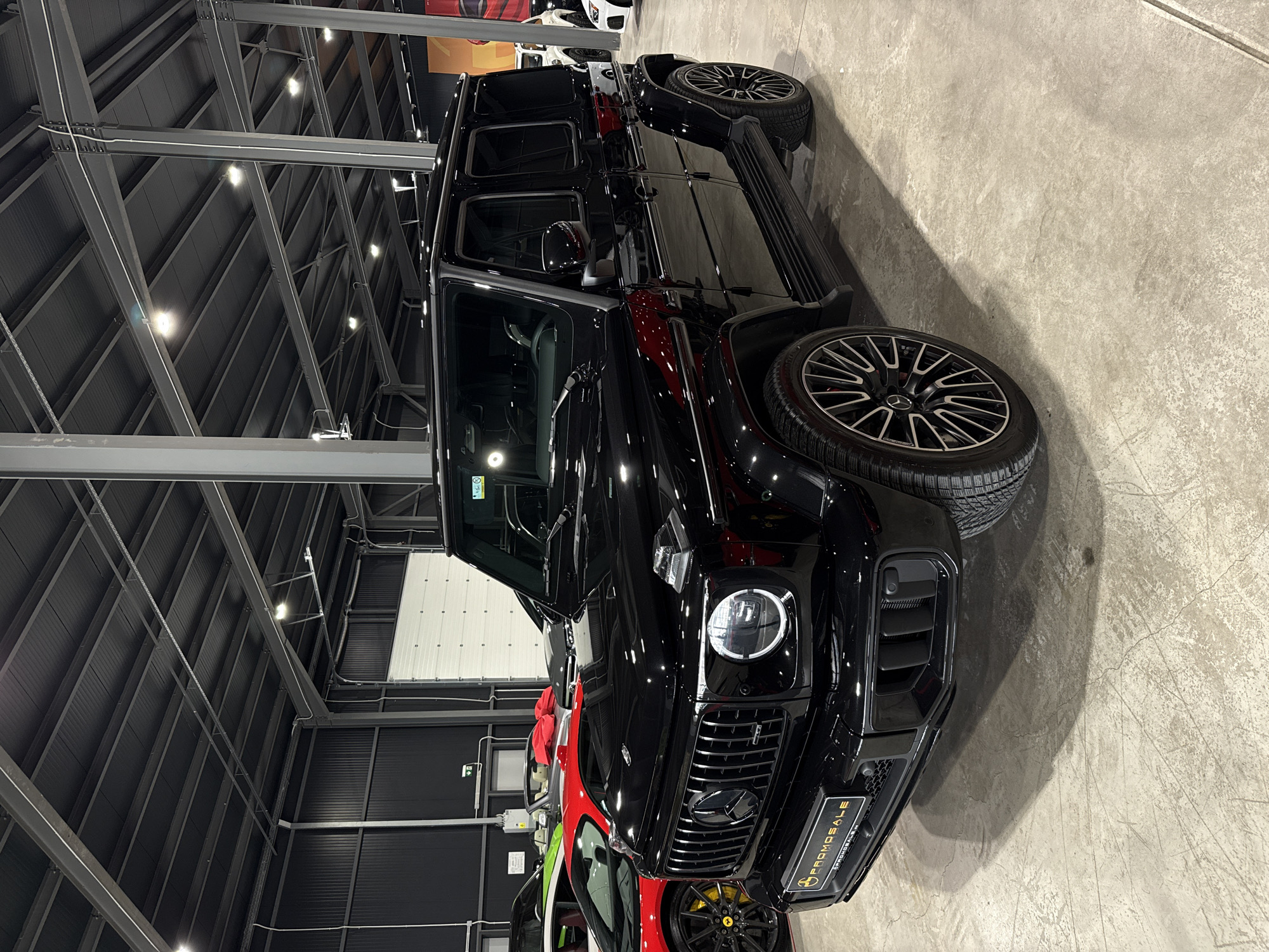 The image shows a black Mercedes-Benz G-Class SUV parked indoors. The vehicle is positioned in what appears to be a dealership or showroom, evidenced by the presence of other vehicles nearby and the building's interior design. The setting has a concrete floor and an industrial-style roof with visible metal beams and lighting fixtures. A promotional sign is visible on the car's front bumper. The specific location in Plovdiv, Bulgaria cannot be determined from the image.