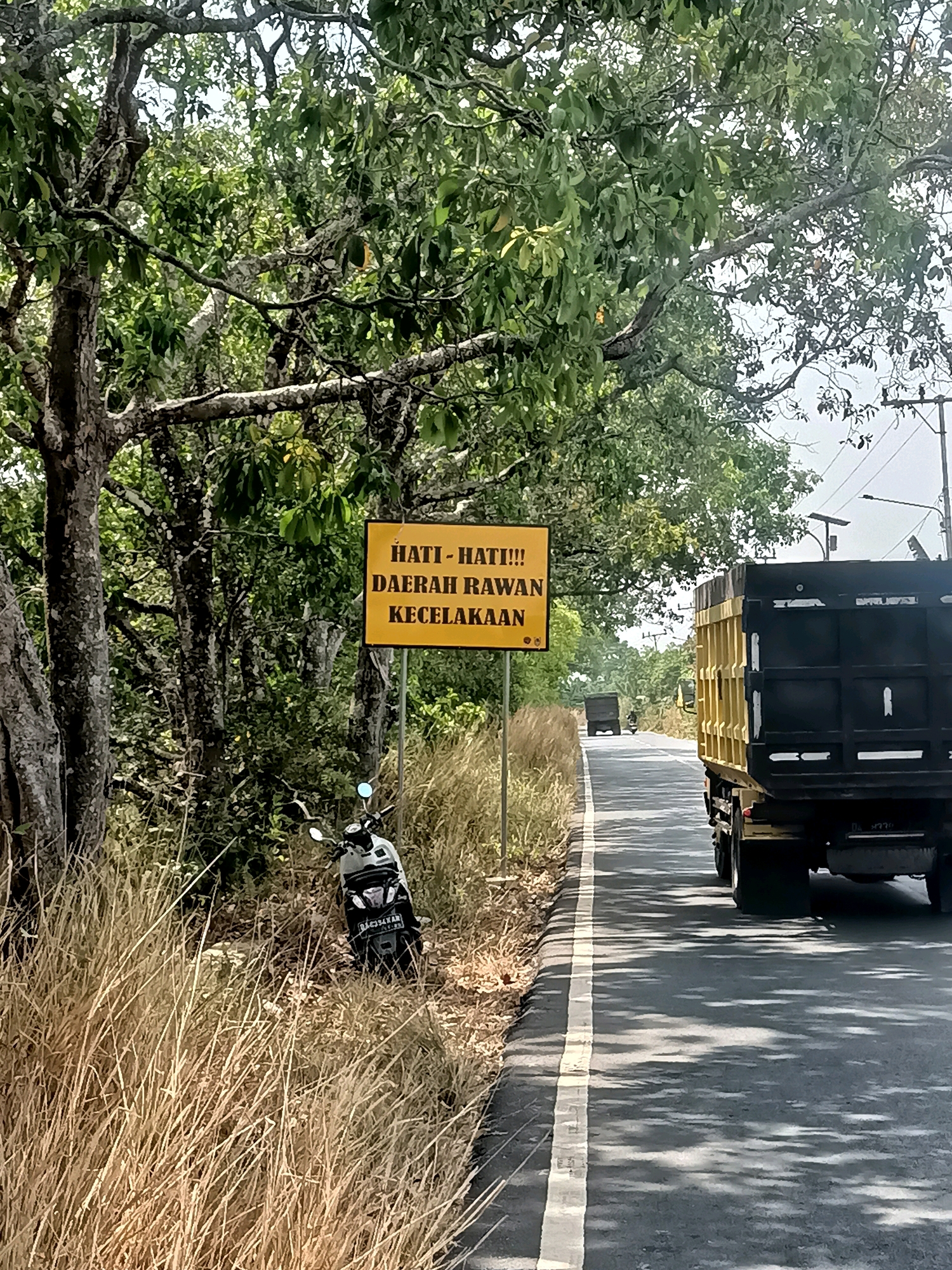 In Sungai Rutas Hulu, Indonesia, a yellow sign with black text reads "HATI - HATI!!! DAERAH RAWAN KECELAKAAN," warning of a dangerous accident-prone area. The sign is positioned beside a road lined with dry grass and trees. A parked motorcycle, likely a scooter, is visible next to the sign. A large yellow and black truck travels down the asphalt road, casting shadows from overhead trees onto the pavement. Another truck is visible further down the road in the distance. The scene appears to be a rural or semi-rural roadside during daylight.
