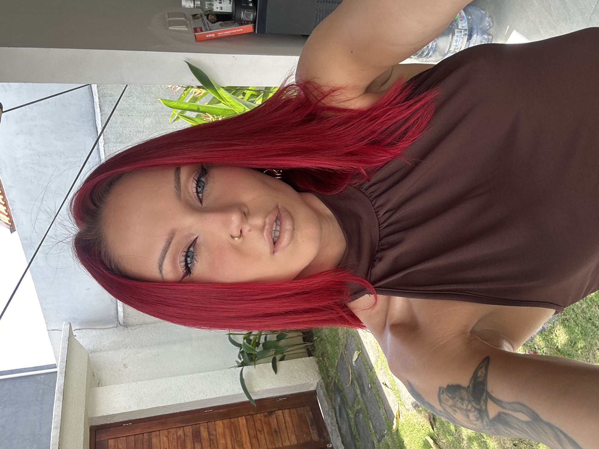 A selfie taken in North Kuta, Indonesia, depicts a person with vibrant red hair and blue eyes, wearing a brown top. The subject has fair skin, noticeable eye makeup, and a nose ring. A tattoo is visible on their arm. The background shows a building with white walls, a wooden door, greenery, and a stone-paved path. In the distance, there are plants and a visible water bottle and other objects on a shelf. The image appears to be taken during daylight.