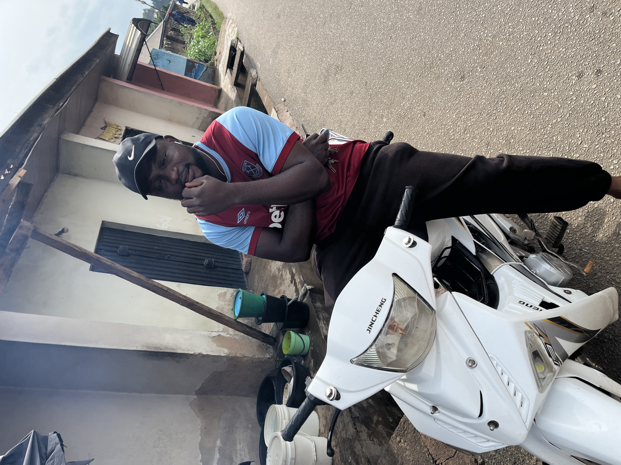 A Black male wearing a Nike cap, a red and blue West Ham United jersey, and dark pants sits on a white scooter. He has his arms crossed over his chest, and his left hand is touching his face. The man is smiling, with his eyes closed. He is in a relaxed pose. The scooter has "JINCHENG" written on the front fairing. In the background, a building with a corrugated metal roof is visible. There are also some buckets and other objects on the ground near the building. The scene appears to be an outdoor setting in a residential area. The time of day is unclear, but the lighting suggests it is daytime. The weather appears to be clear and sunny. The location cues, such as the building style and the scooter model, are consistent with an urban or semi-urban environment in Nigeria. The overall mood of the image is casual and friendly.