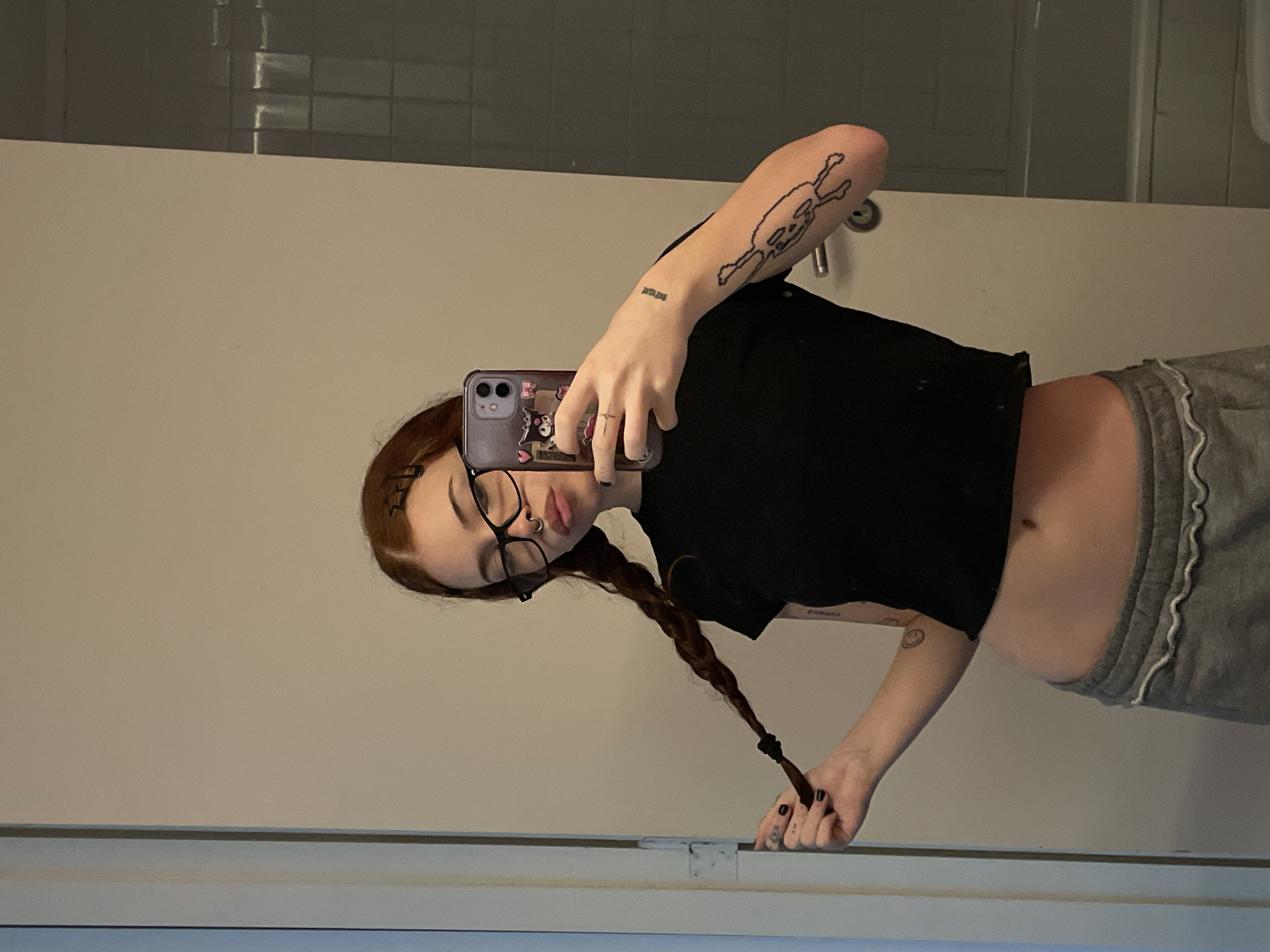 The scene depicts an indoor mirror selfie of a young female subject. The location is an interior space, consistent with a private residence or facility in Buenos Aires, Argentina. The subject has reddish-brown hair styled in two braids, secured with black hair ties; her left hand holds the end of one braid. She wears black-framed glasses, a septum nose piercing, and a small ring on her right ring finger. Her attire includes a black crop top, exposing her midriff, and grey sweatpants with white ruffle trim along the waistband. Visible tattoos consist of a large, outlined skeletal or humanoid figure on her right bicep/forearm, small text on her inner right forearm, and a small circular design on her left bicep. Her fingernails are painted dark. She holds an iPhone, featuring a purple case adorned with cartoon characters, in her right hand, positioned to capture her reflection. Her lips are slightly pursed. The background shows a light beige wall, a vertical white doorframe on the left, and a section of a bathroom on the right with grey tiled walls, a silver lever-style door handle, and a partial view of a white bathtub or shower basin. The lighting is artificial and warm.