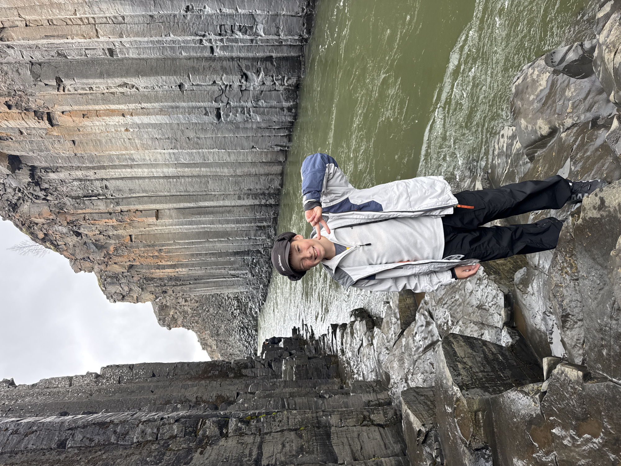 The image, when correctly oriented, depicts a young person standing on the rocky bank of a river, surrounded by impressive columnar basalt formations. The person, appearing to be a male child or pre-teen, is in the mid-ground on the right, smiling and making a peace sign with their left hand. They are dressed in a light grey and navy blue jacket, black trousers, and a dark baseball cap with light text on the front, possibly "Hatlands."
The scene is a deep gorge or canyon. Towering, dark grey basalt columns form the steep, geometric walls on both sides of the river, creating a visually striking natural environment. The rocks in the foreground and along the river's edge are visibly wet and glistening, suggesting recent rain or moisture. The river itself is a murky green color with visible currents and ripples. The sky, visible in the upper left, is overcast and grey, indicating a cloudy daytime. The overall impression is one of rugged natural beauty, with the person's joyful expression adding a sense of awe and enjoyment of the unique geological landscape. The distinct hexagonal basalt columns are characteristic of locations such as Stuðlagil Canyon in Iceland.