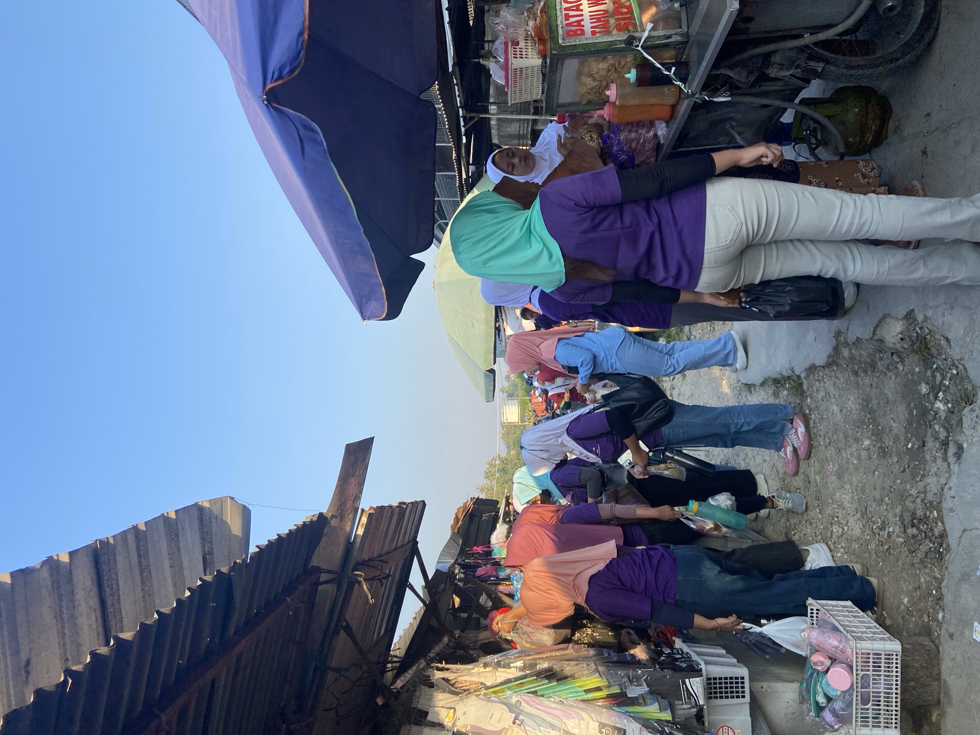 A street scene in Tanjungrejo, Indonesia, captures a bustling market atmosphere. The image, taken during daylight hours with clear blue skies, features several individuals engaged in shopping and vendor interactions.
On the left side, a stall with a corrugated metal roof displays various goods, including clothing items packaged in plastic. A woman wearing a light pink hijab and a purple shirt stands near this stall, examining the merchandise.
Further into the scene, a group of women, most wearing purple shirts and headscarves in various colors (light pink, white, and teal), are gathered. One woman in a white hijab and a white top appears to be a vendor, standing behind a cart filled with food items and condiments. A green gas cylinder is visible beneath the cart. A sign on the cart reads "BATAG TAHU WA SIOS".
Several people are holding shopping bags, suggesting active commerce. The ground is a mixture of dirt and gravel. In the background, a large blue umbrella partially shades a section of the market, and other market stalls and people are visible, indicating a lively marketplace environment. The overall scene depicts a typical day at an outdoor market in Indonesia.