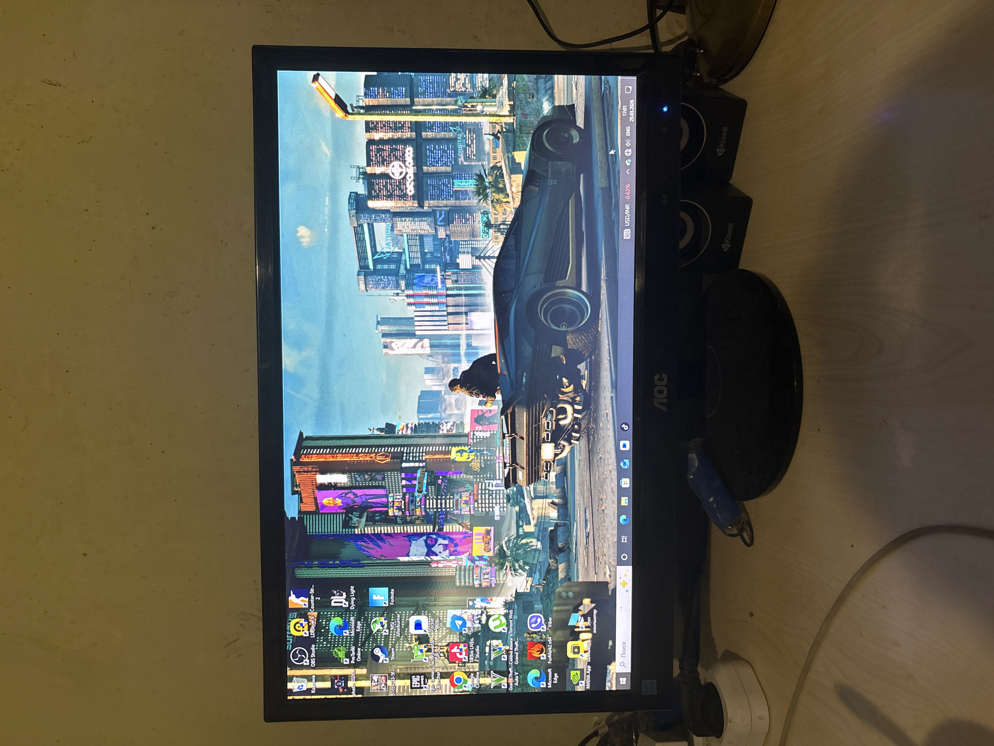 The image shows a computer monitor displaying a wallpaper featuring a futuristic cityscape, likely inspired by the game Cyberpunk 2077. The monitor is an AOC brand and has visible icons on the desktop, including software like OBS Studio, a game emulator LDPJayer, Counter-Strike, Dying Light, Fortnite, Grand Theft Auto V, Microsoft Edge, and FurMark2 x64. The taskbar shows the current date and time (29.03.2026, 17:01), language settings (ENG), and currency (USD/INR). Additionally, there are Kisonli speakers adjacent to the monitor. The surrounding environment suggests an indoor setting with a wall and desk. The image does not show any location in Yerevan, Armenia.