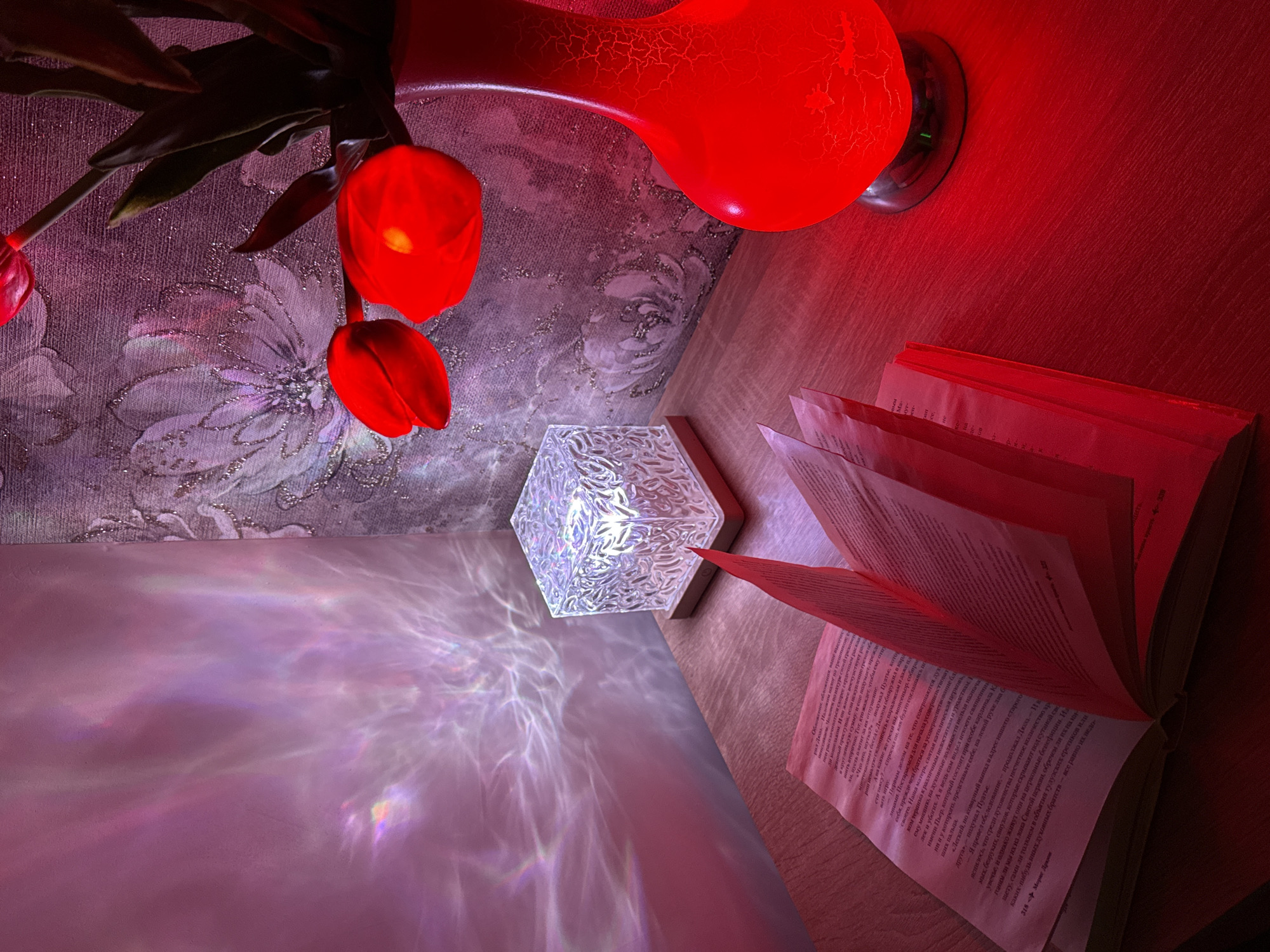 The image depicts an indoor scene featuring a floral arrangement in a red vase, a decorative hexagonal lamp, and an open book. The vase holds red tulips with green leaves. The lamp is white and casts a patterned light onto a nearby surface. The open book lies on a wooden surface. The scene is bathed in a predominantly red light, with highlights from the lamp. The wallpaper features a floral pattern.