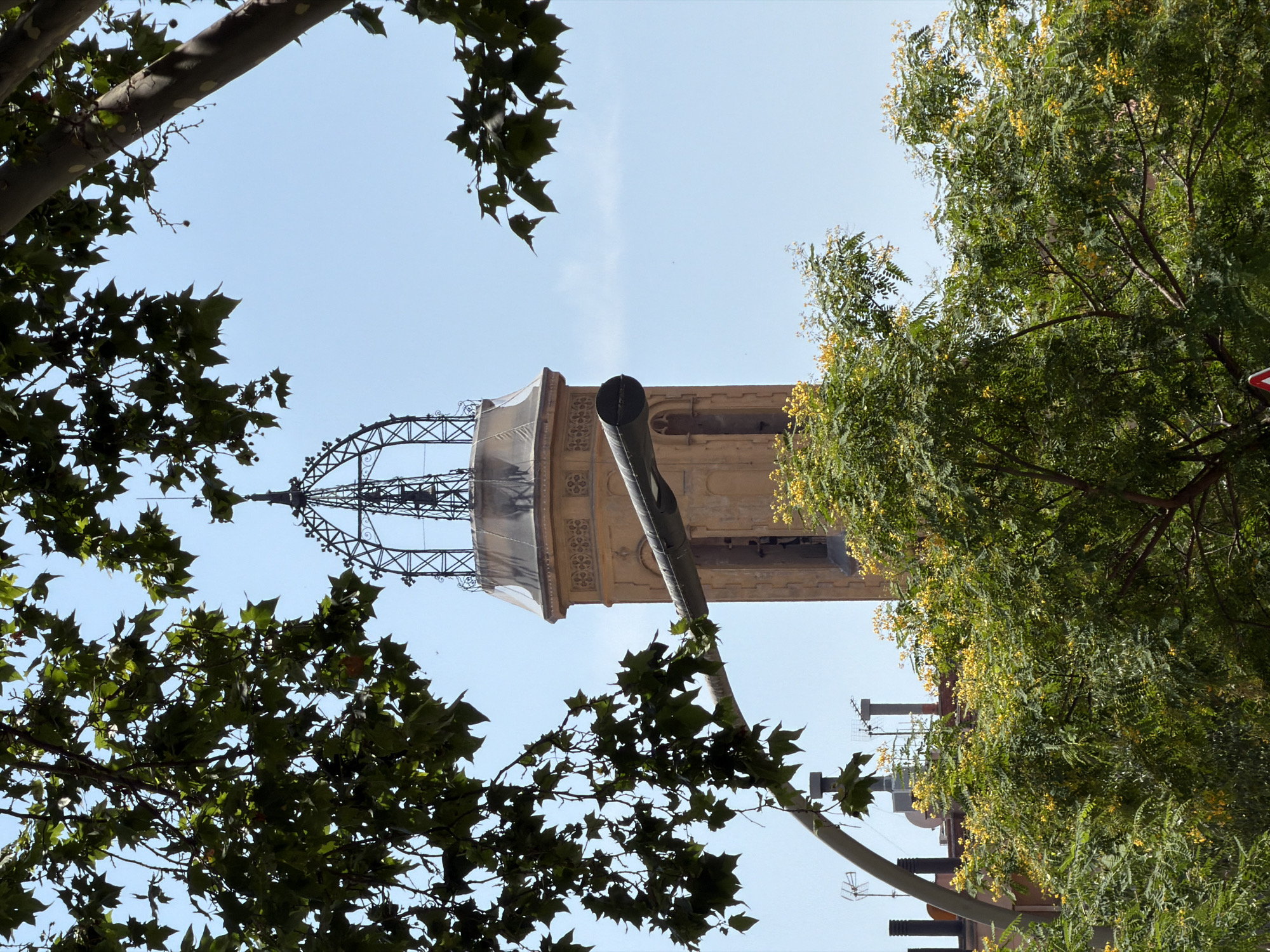 The image shows a view through the branches of trees towards a tower in Barcelona, Spain. The tower is light brown with decorative patterns and a pointed, wrought-iron spire. In front of the tower, a dark, cylindrical object, possibly a lamp or conduit, extends horizontally across the frame. Lush green trees with clusters of small yellow flowers are visible in the foreground and middle ground, obscuring parts of the lower portion of the tower and surrounding buildings. The sky is clear and light blue.
