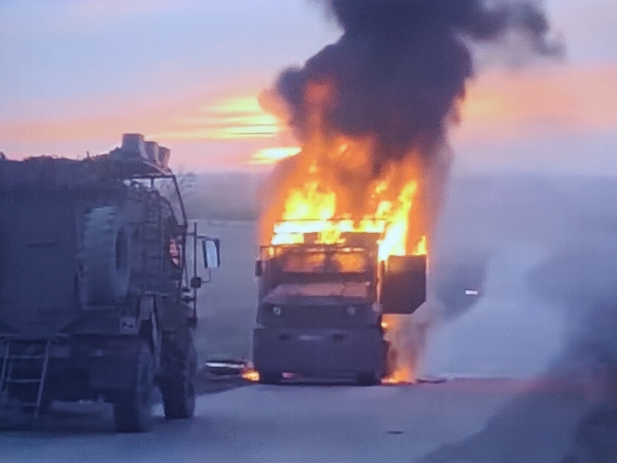 The image depicts a scene at dusk on a road, likely in the vicinity of Molenbeek-Saint-Jean - Sint-Jans-Molenbeek, Belgium. In the foreground, on the left, is a large military-style truck with a spare tire mounted on its side. To the right of this truck, another, smaller truck is engulfed in flames. The fire is intense, producing thick black smoke that billows upwards into the hazy sky. The sky itself shows the colors of sunset, with hues of orange, pink, and purple. The ground appears to be asphalt. The overall impression is one of destruction and a dramatic, possibly post-incident, environment. There are no people visible in the image.