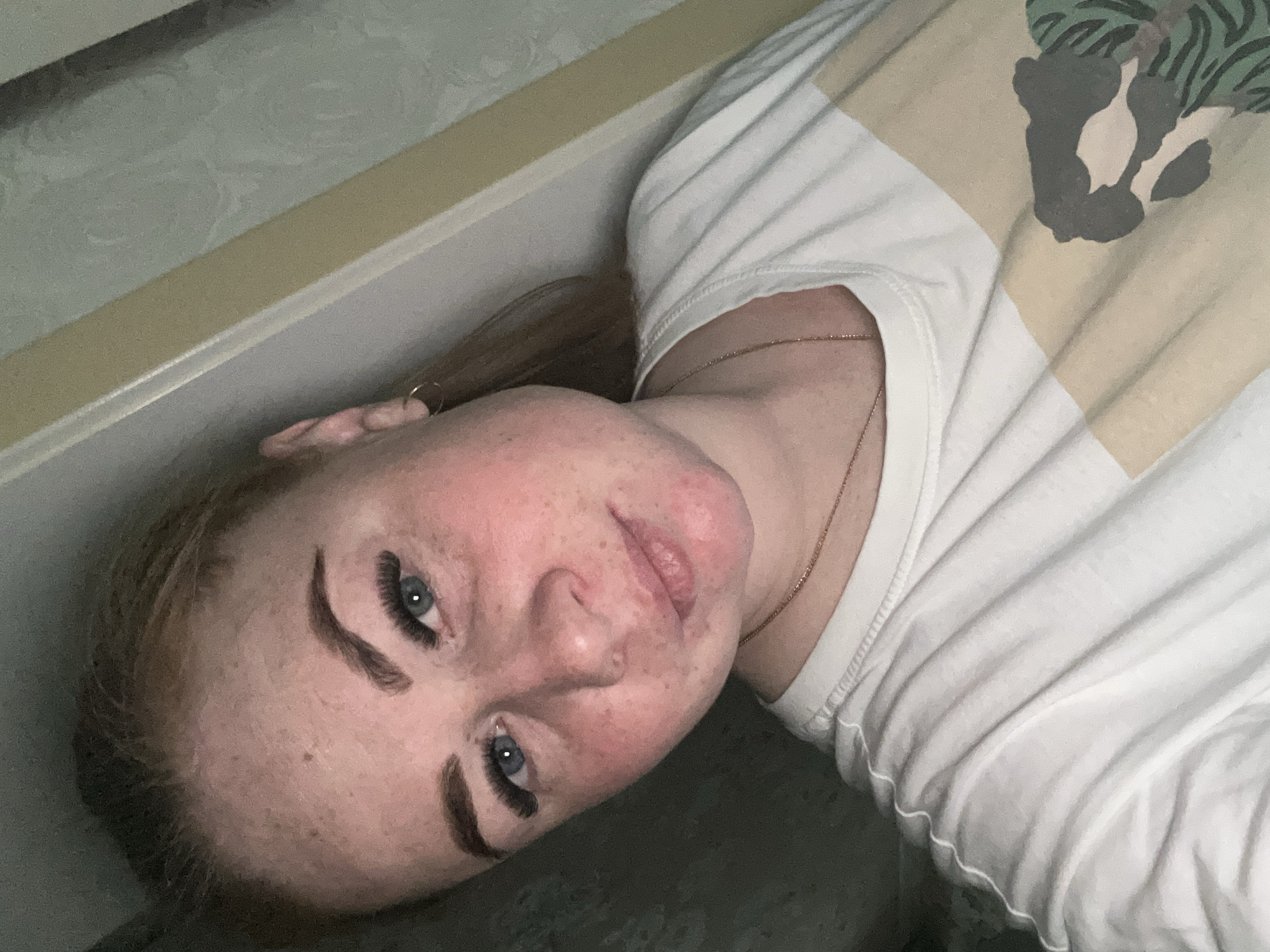 A fair-skinned woman with freckles, wearing a white t-shirt with a printed design, is lying down looking at the camera. She has blonde hair and blue eyes and wears an earring. The background includes a textured wall. The location cannot be determined from the given image.