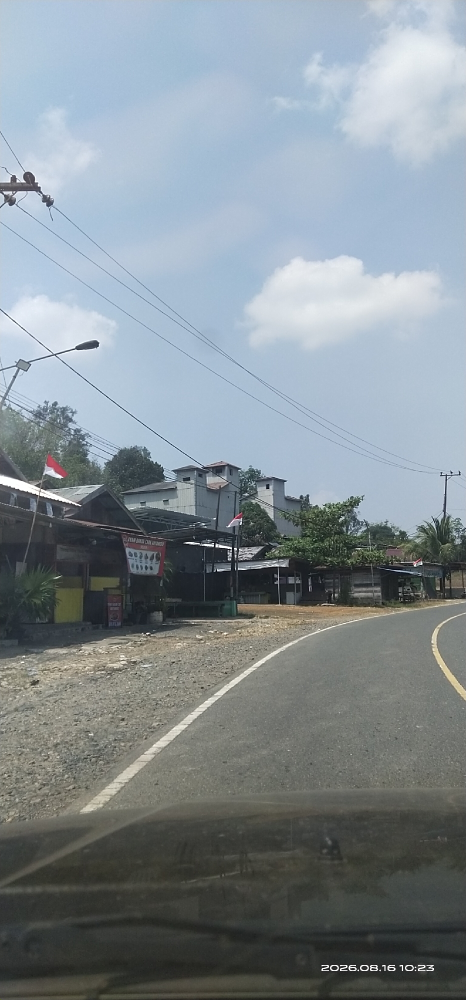 A road curves through a semi-rural area under a bright, partly cloudy sky. The road is asphalt with a yellow line on the right edge and a white line on the left. To the left of the road are several small buildings, possibly shops or stalls, with corrugated metal roofs. Some of these buildings have signs and flags. Indonesian flags are visible on two of the buildings. Behind these structures are trees and larger, multi-story buildings that appear to be factories or industrial facilities. Power lines and a street light are also present. The foreground shows the hood and windshield of a vehicle, reflecting the scene ahead. A date and time stamp, "2026.08.16 10:23", is in the bottom right corner. The scene appears to be a roadside environment in an Indonesian town or city, likely Puri based on the information provided.