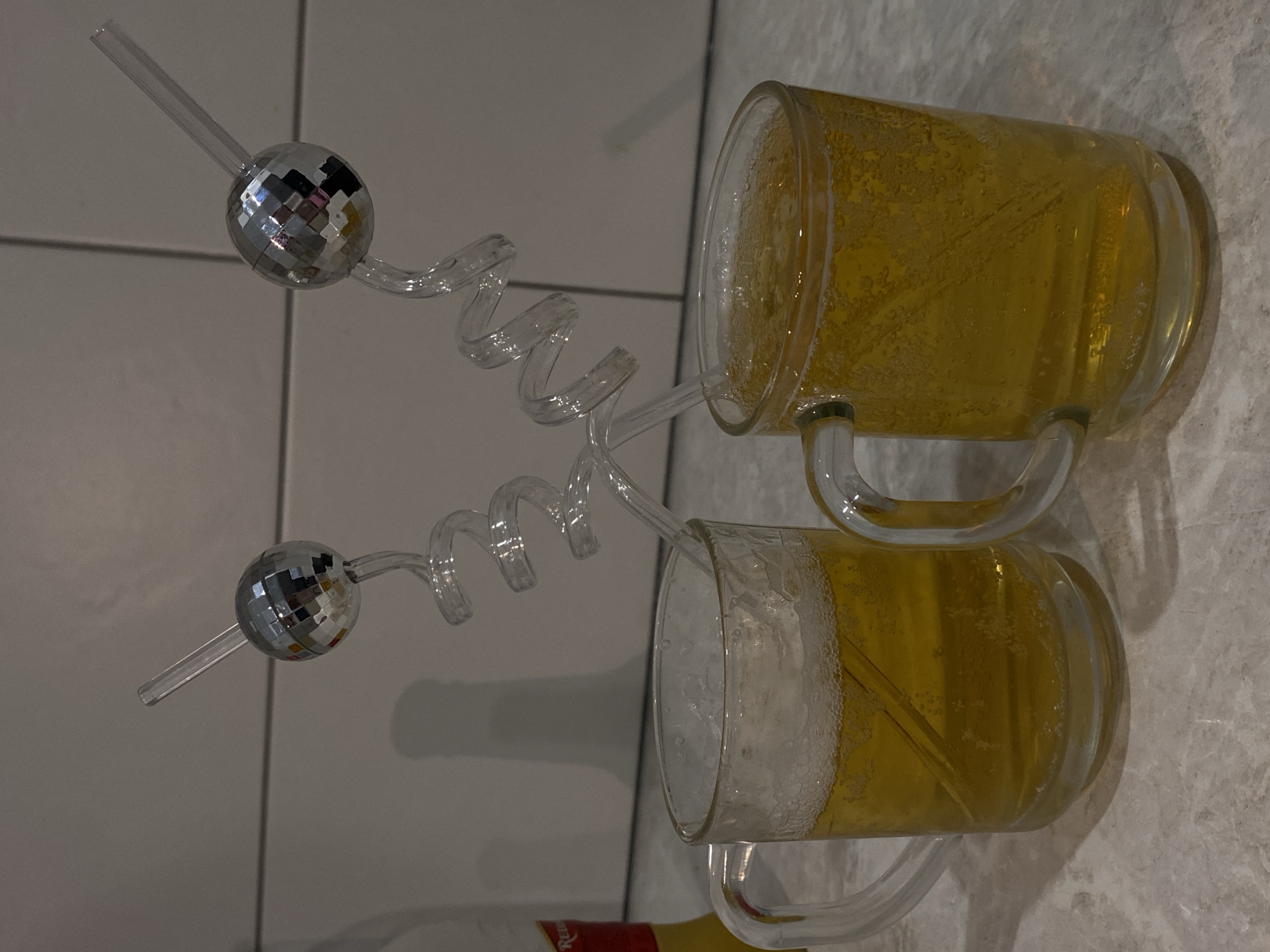 The image displays two clear glass beer mugs, positioned horizontally in the frame, each filled with a light golden, bubbly liquid topped with a layer of white foam. Both mugs feature a distinctive clear plastic straw with a spiral design, each topped with a small, reflective disco ball ornament. The liquid inside the mugs shows numerous small bubbles adhering to the glass.
The mugs are placed on a light-colored, subtly textured surface, possibly a tiled countertop or table. A light grey wall or background with visible vertical and horizontal lines is behind them. In the bottom left corner, partially obscured, is a bottle with a red and yellow label, on which the text "Red" is faintly visible.
The scene appears to be an indoor setting, likely under artificial or ambient light, as evidenced by the shadows cast by the mugs and straws. There are no people visible, and the items are arranged as if prepared for consumption. No specific time of day or weather conditions can be determined from the image.