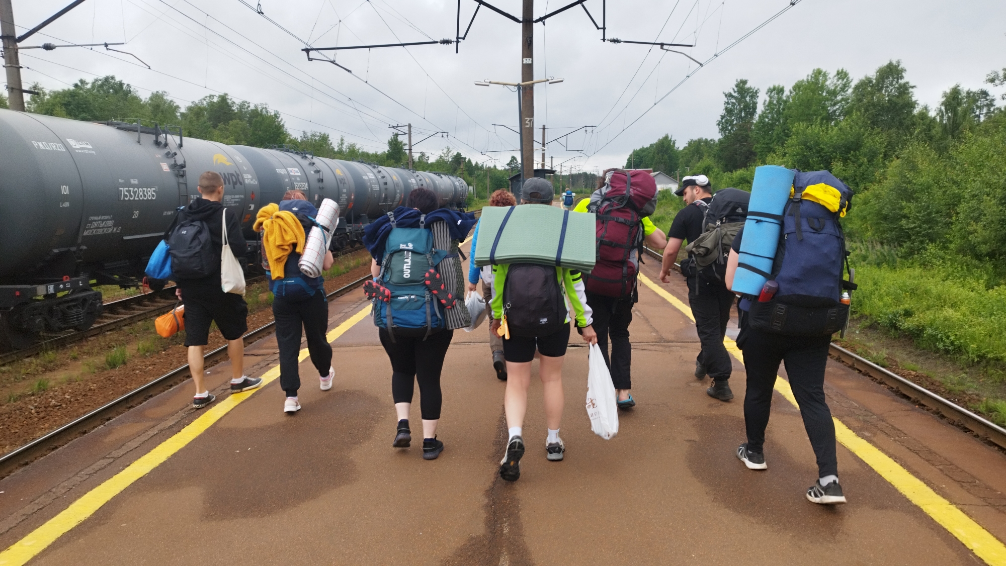 A group of eight individuals, each carrying large backpacks, are walking away from the camera on a train platform in Orekhovo, Russia. The platform is paved with asphalt and has a yellow line marking the edge. To the left of the platform are train tracks and a long, dark-colored train composed of multiple carriages. The train carriages appear to be tankers, with visible markings and numbers, including "75328385". Overhead, power lines for the train are visible. The background consists of dense green trees and foliage, with a cloudy sky overhead. The individuals are dressed in casual clothing suitable for travel or outdoor activities. Some are wearing hats and headphones. The backpacks are of various colors and sizes, some with sleeping mats or other camping gear attached. Some individuals are also carrying bags or bottles. The scene suggests a departure or arrival of a group on a train journey.