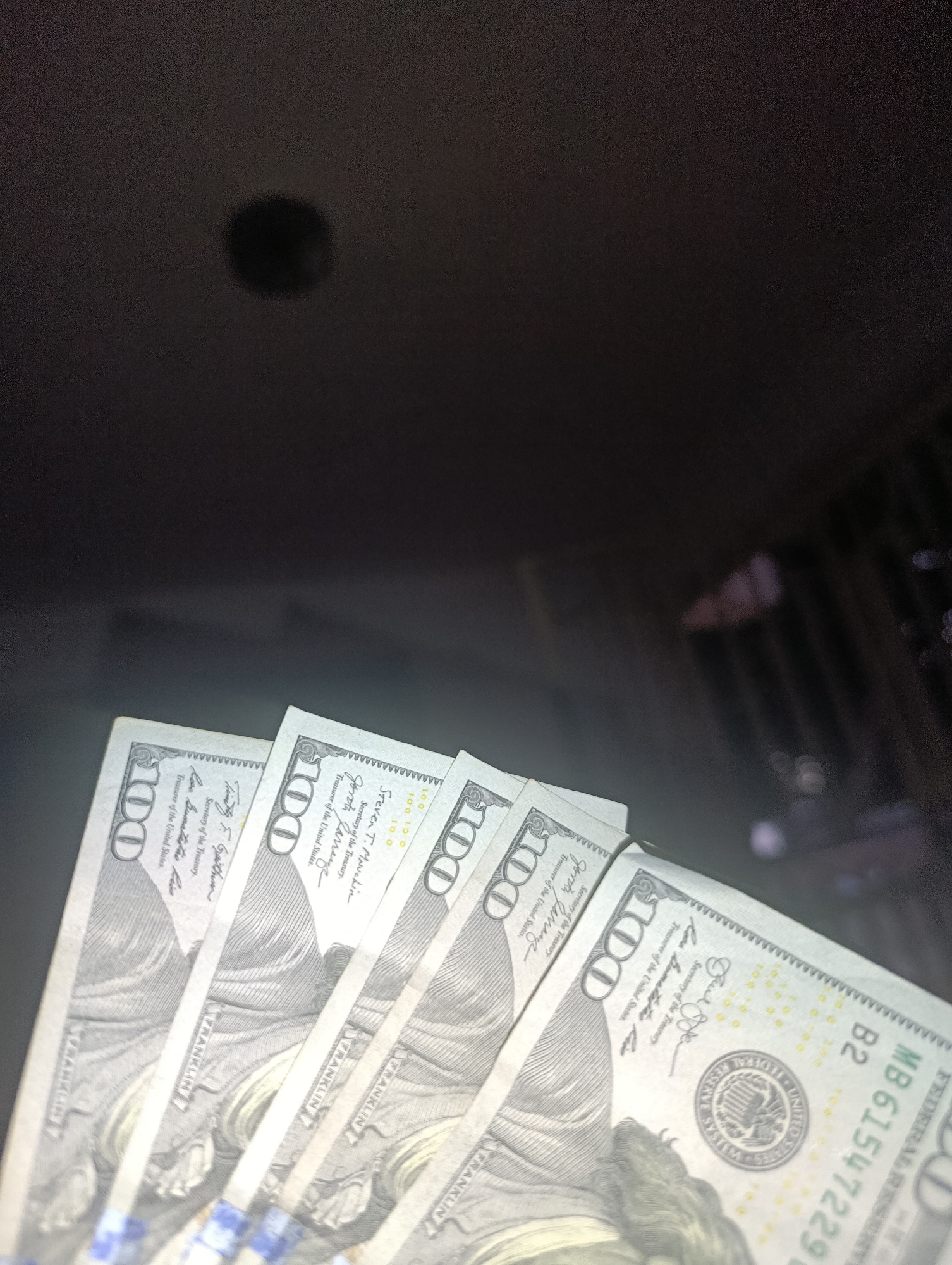 A close-up shot captures several US $100 bills fanned out against a dark, indistinct background. The bills are held or laid out in a slight overlap, creating a diagonal composition across the lower half of the frame. The lighting is dim, emphasizing the details of the currency while leaving the environment mostly obscured, save for a blurry, dark circular object near the top center and some faint, out-of-focus elements on the far right.
Each bill prominently features the denomination "100" and the image of Benjamin Franklin. Visible text includes "FEDERAL RESERVE NOTE," "UNITED STATES OF AMERICA," and "FRANKLIN." On at least one bill, the signatures of "Steven T. Mnuchin" (Secretary of the Treasury) and "Jovita Carranza" (Treasurer of the United States) are clearly readable. A serial number, "MB 61547229 B2," is visible on the rightmost bill, along with the "FEDERAL RESERVE SYSTEM" seal. The overall scene suggests a focus on the monetary value, possibly in the context of a transaction or display of wealth in Juan Griego, Venezuela.
