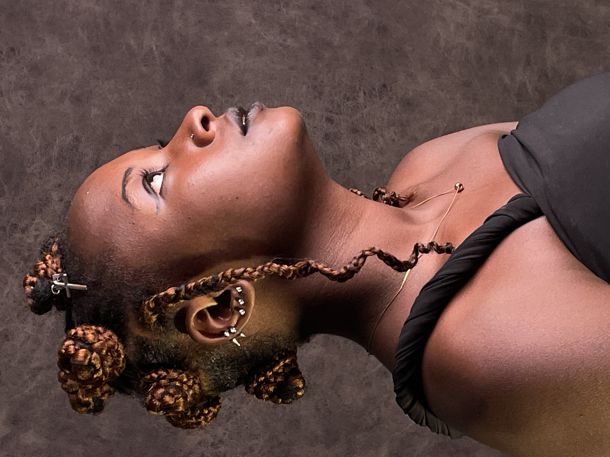 This is an artful, close-up portrait featuring a person with dark skin, likely a woman, her head tilted back and angled slightly to the left. The subject is looking upwards with a contemplative expression. Her face is accentuated by dark, glossy lips and well-defined eyebrows. She wears a small stud nose piercing and multiple ear piercings adorned with silver-toned jewelry, including a cluster of studs on the upper ear and a spiky earring on the lobe.
Her hair is intricately styled in braids, some of which are gathered into large, decorative knots around her head, secured with small metallic accessories. A long, coiled braid extends from behind her ear down towards her shoulder. She is dressed in a black garment, possibly a top or dress, and a delicate gold chain necklace with a small pendant rests on her collarbone. The background is a plain, dark, textured surface, providing a stark contrast that highlights the subject. The soft lighting emphasizes her features and the details of her hairstyle and accessories. The portrait conveys a sense of introspection or aspiration. No text is visible in the image. The location context provided is Barakin Akawo, Nigeria.