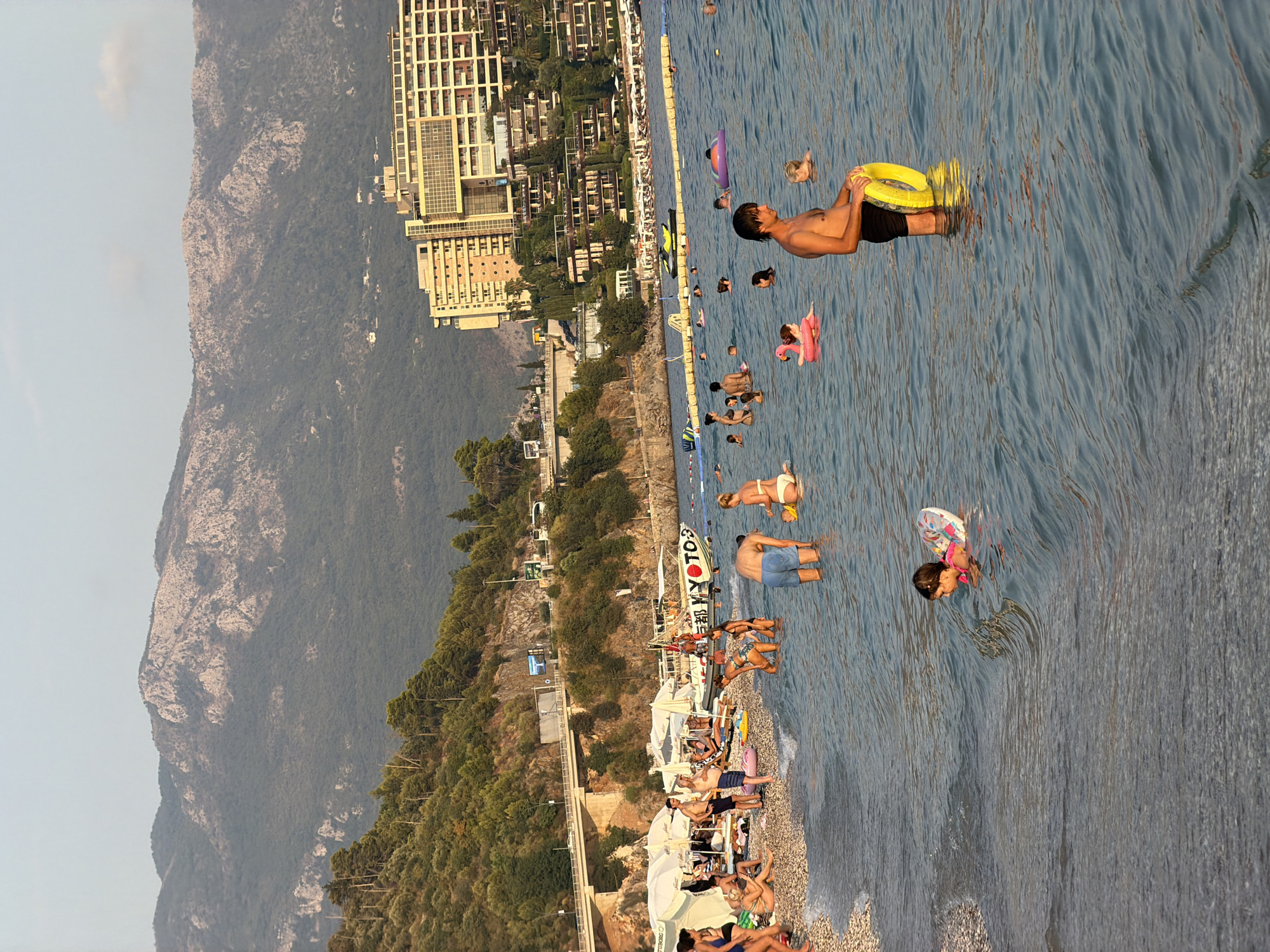 The image depicts a crowded beach scene in Budva, Montenegro, with numerous people enjoying the water and the shore. In the foreground, a man stands in the shallow water holding a yellow inflatable ring, while other individuals are visible swimming and floating further out. A young child wearing a pink and white inflatable ring is also in the water. The beach itself is covered in pebbles, and several white umbrellas provide shade for sunbathers. In the background, a large, modern hotel complex is visible, built into the hillside. Above the hotel, steep, green-covered mountains rise, with a clear sky overhead. A road and bridge are also visible along the coastline. The overall impression is a busy, sunny day at a popular tourist destination.